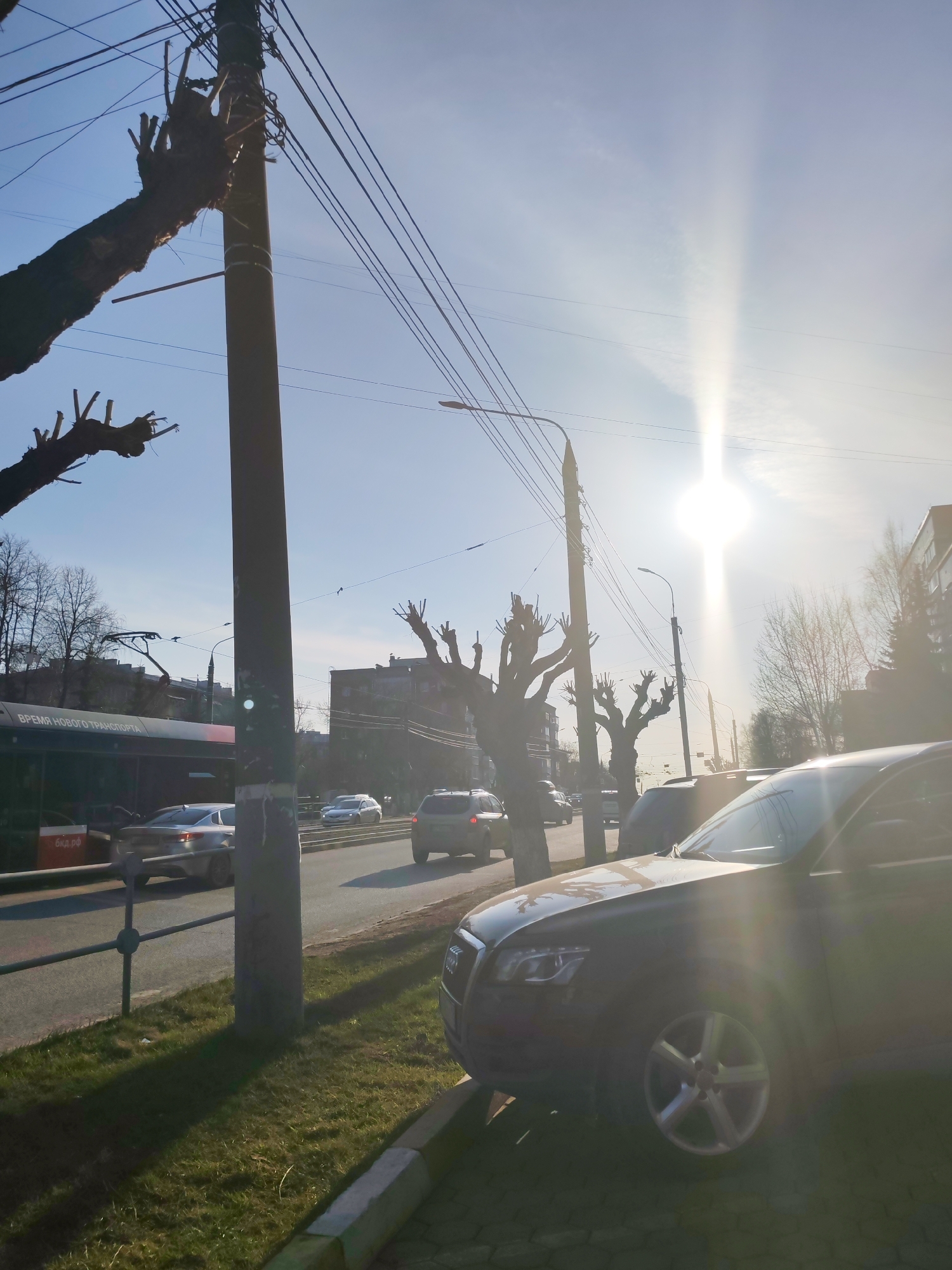 The image captures a street scene in Izhevsk, Russia, on a sunny day. A dark-colored Audi SUV is parked on the right side of the frame, partially obscuring the view. In the background, several cars are driving on the road. On the left, a bus stop with a sign written in Russian is visible. Bare trees line the street, with utility poles and wires overhead. Buildings are present in the background, but they appear somewhat blurred due to the distance and sunlight. The sun is high in the sky, creating a bright glare. Grass and a curb are visible in the foreground.