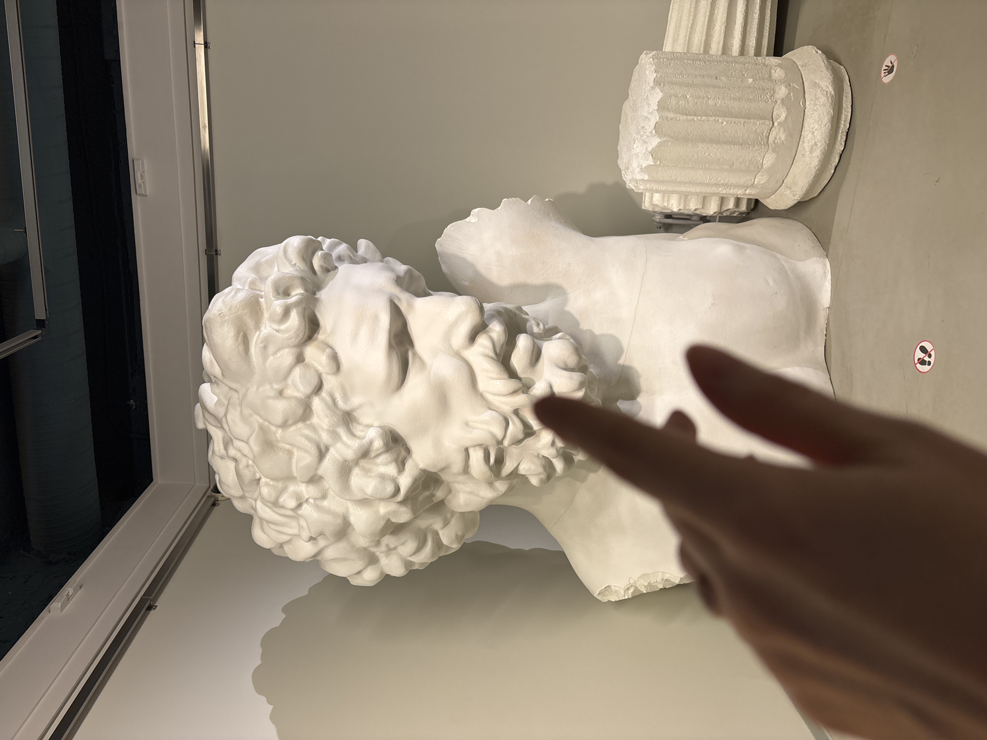 An indoor, medium-shot image depicts a white, plaster cast of a man's head and torso with a column capital behind, possibly of Hellenistic origin. The subject lies on a light beige surface inside a museum, next to a window. A hand, likely belonging to a visitor, is pointing towards the head of the statue. Two round stickers with a crossed-out foot and a hand are visible on the wall, indicating that touching or stepping over the statue is prohibited. The location is possibly in Saint Petersburg, Russia.