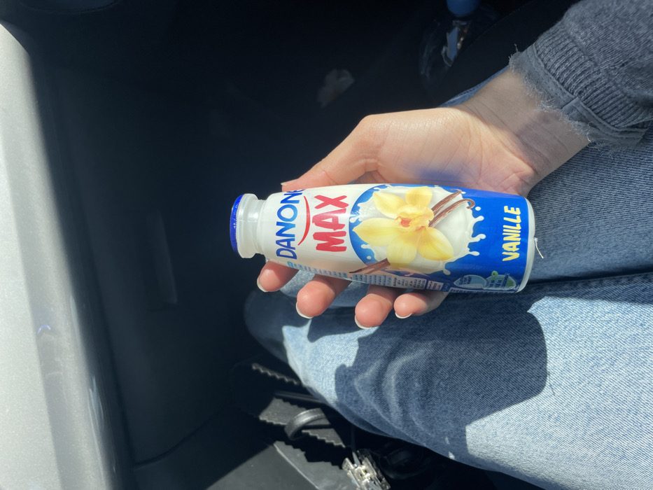The image depicts a close-up view of a person's left hand holding a white plastic bottle of Danone MAX Vanille yogurt drink. The hand exhibits manicured fingernails with white tips. The person is wearing light blue denim jeans and a grey long-sleeved top with a visibly frayed cuff on the right wrist. The Danone bottle has a blue screw cap and a label featuring a yellow vanilla flower and a vanilla bean against a white liquid splash on a blue background, with "DANONE" in blue, "MAX" in red, and "VANILLE" in yellow text. The setting appears to be inside a vehicle, indicated by dark interior surfaces and a lighter grey panel visible in the upper left. Strong sunlight casts clear shadows within the vehicle's interior. A dark bag or purse with a silver buckle is partially visible to the left of the person's leg. This scene is located in Skhirat, Morocco.