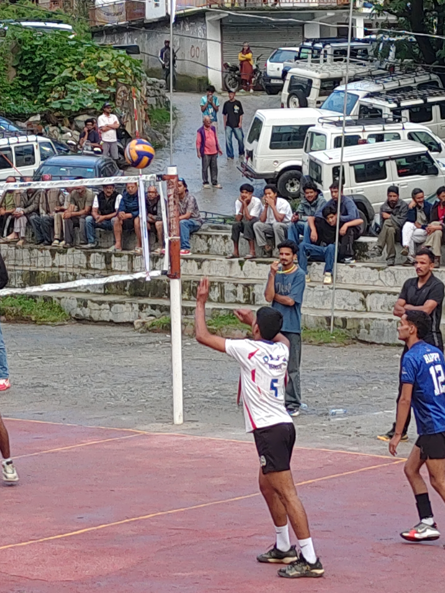 A group of young men are playing volleyball on a red court with yellow lines, with a backdrop of a village and steep hills. One man in a white jersey with red trim and the number 5 is serving the ball. Another man in a blue jersey with the number 12 is on the opposing team. Spectators are sitting on stone bleachers in the background, some watching the game, others talking. A volleyball net is stretched across the court. The environment appears to be a small village, possibly Burwa, India, with traditional housing and vehicles parked on a street. The weather seems to be overcast.