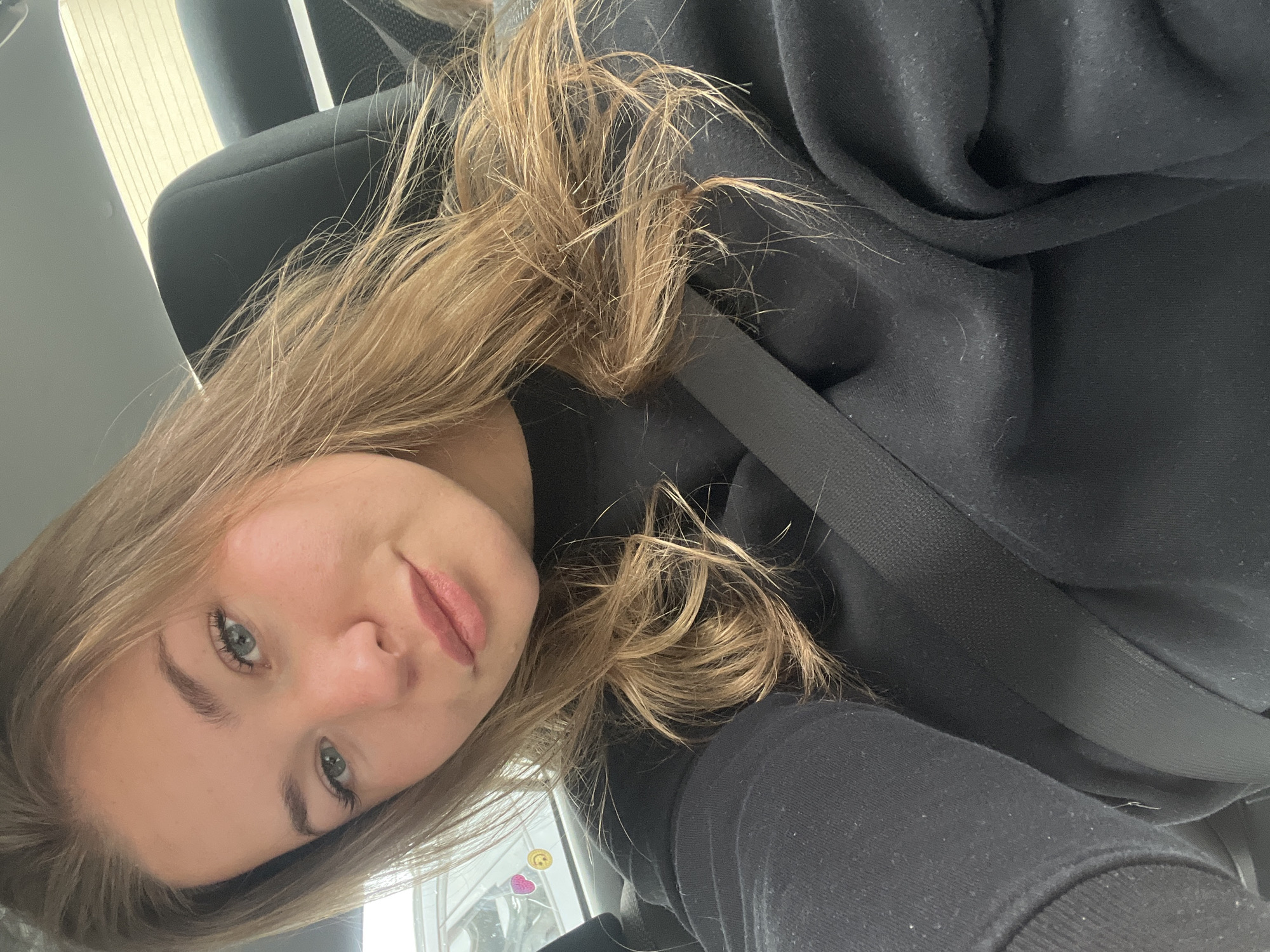A fair-skinned female with light brown hair and blue eyes is positioned inside a vehicle, facing the camera at an angle. She is wearing a black hoodie, and a seatbelt is fastened across her chest. The vehicle's interior and a glimpse of the outdoors are visible. The location is not discernible as being Budva, Montenegro from the visual information provided.