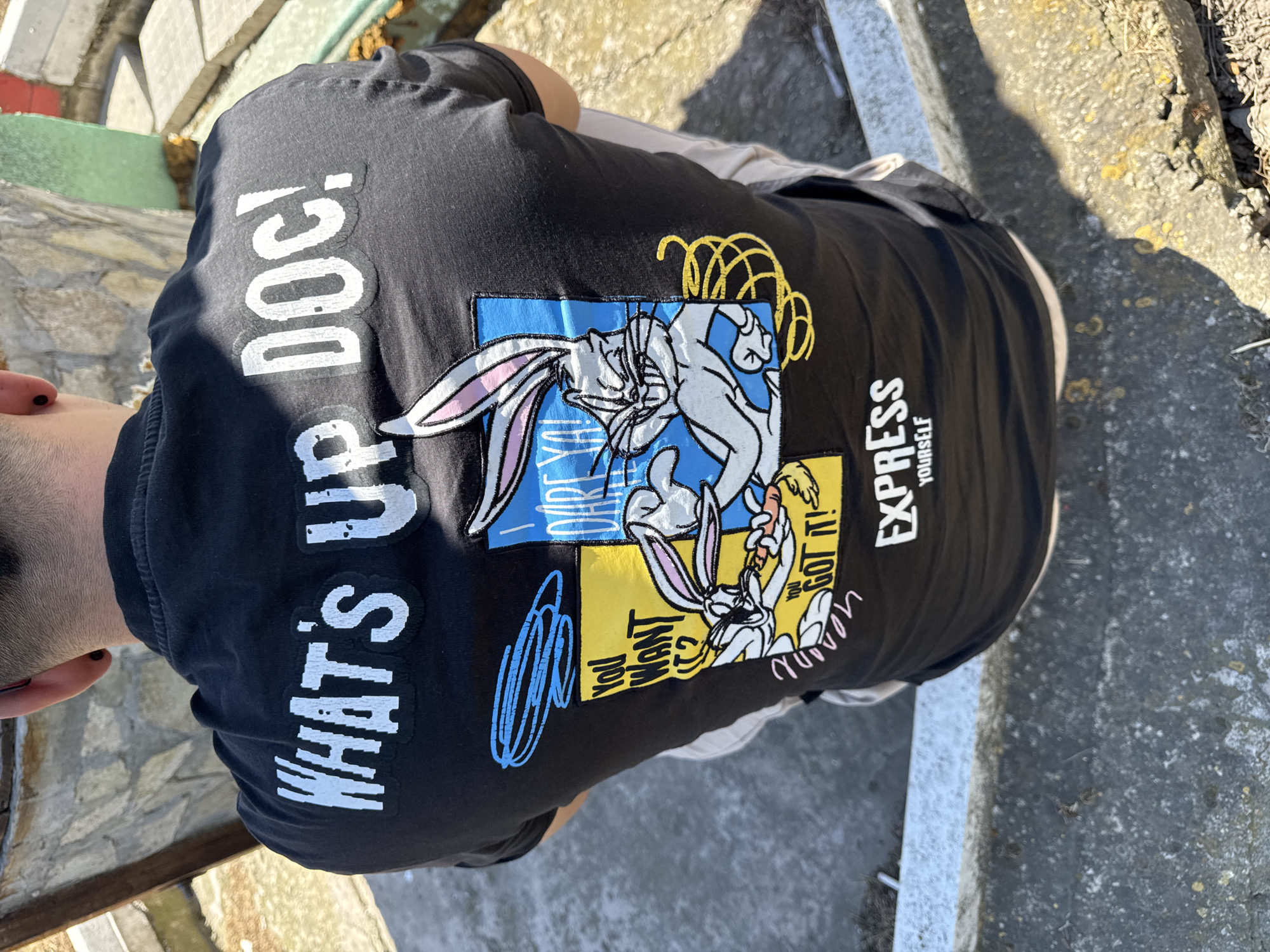 A person is wearing a black T-shirt with a Bugs Bunny design and the phrase "WHAT'S UP DOC!" in white lettering. The design includes Bugs Bunny images in blue and yellow squares. The person is standing outdoors on a concrete surface with textured stone or mosaic-like sections. The location is in the city of Probuda, Bulgaria.