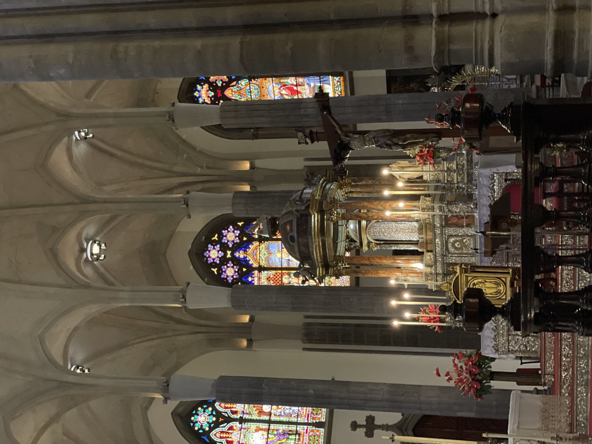 The image displays the grand interior of a church or cathedral in San Cristóbal De La Laguna, Spain. The scene is viewed from an aisle, looking towards the main altar.
**Subjects:** The dominant architectural features include tall, fluted stone columns with decorative capitals, which support elaborate vaulted ceilings in a light, neutral color. Multiple stained glass windows are visible, including round rose windows with intricate patterns and rectangular ones depicting religious figures and scenes. At the focal point is a highly ornate main altar or retable, featuring rich gold and silver elements. Above the altar stands a prominent crucifix with the figure of Christ, draped with small red and white pennants. A gold-colored structure, possibly a tabernacle or monstrance, is central to the altar. Another religious figure (possibly a statue) is partially visible to the left.
**Activity or event taking place:** No people are visible, and no specific activity or event is ongoing, presenting a quiet and contemplative scene within the sacred space.
**Notable details:** The interior is dimly lit, with light primarily emanating from the vibrant stained glass windows, suggesting it is daytime. Lit candles on the altar contribute to the ambiance, and arrangements of red flowers adorn the altar and a nearby pedestal, adding color to the solemn setting. The inscription "INRI" is clearly visible above the figure of Christ on the crucifix. The grandeur of the architecture and the religious artifacts evoke a sense of reverence and spiritual solemnity.