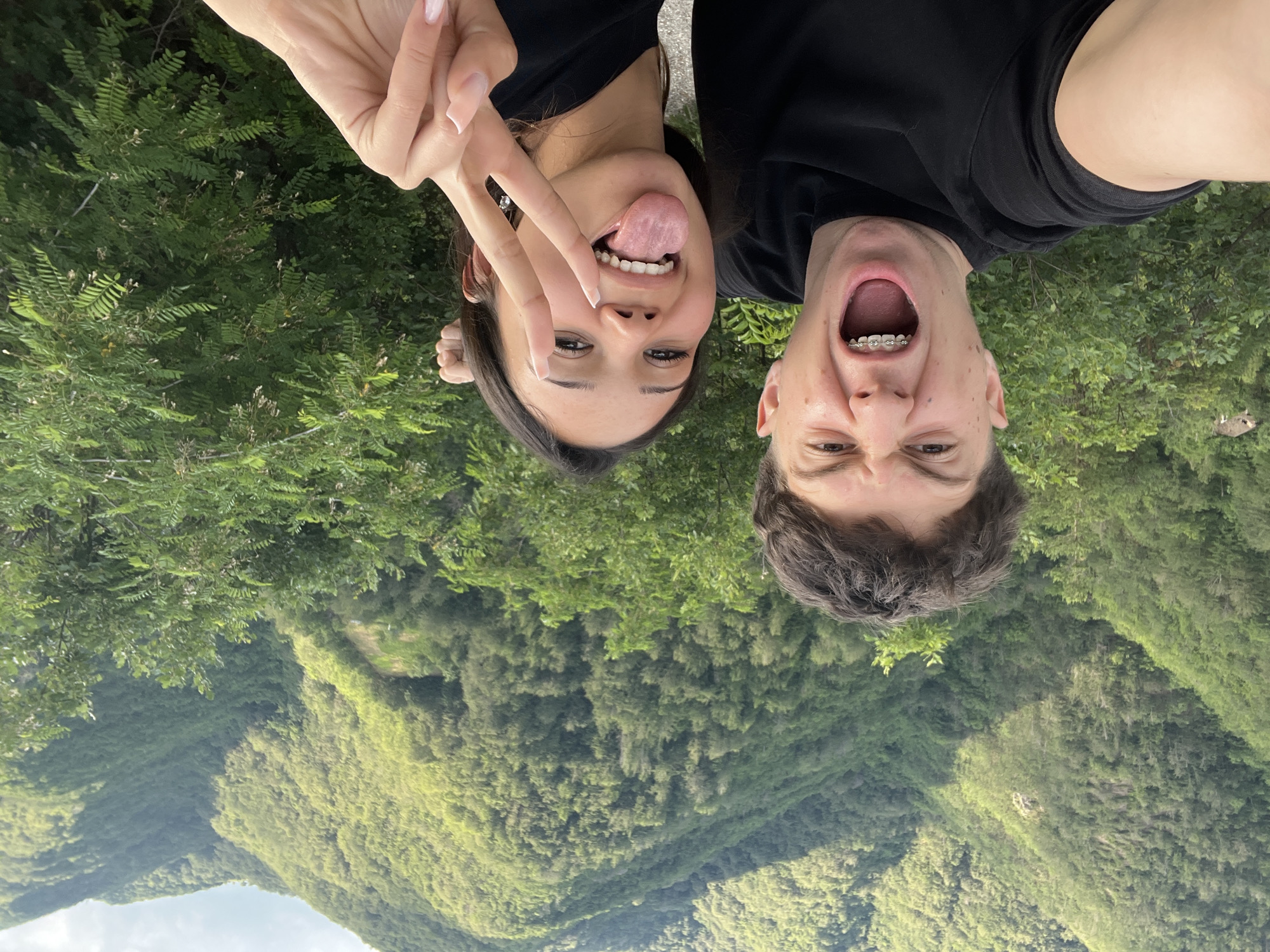 In Pechinska, Bulgaria, a young couple is captured in a selfie with a lush, mountainous backdrop. The man, on the left, has a playful expression, mouth open wide and teeth with braces visible, his right arm extending towards the camera. The woman, on the right, sticks her tongue out playfully and makes a peace sign with her left hand, her right arm also visible. They are both wearing black t-shirts, and she has long, light-colored fingernails. The environment behind them is a dense green forest covering steep hillsides, with a visible path or road in the foreground and a hint of a building on the far left. The sky above is cloudy.