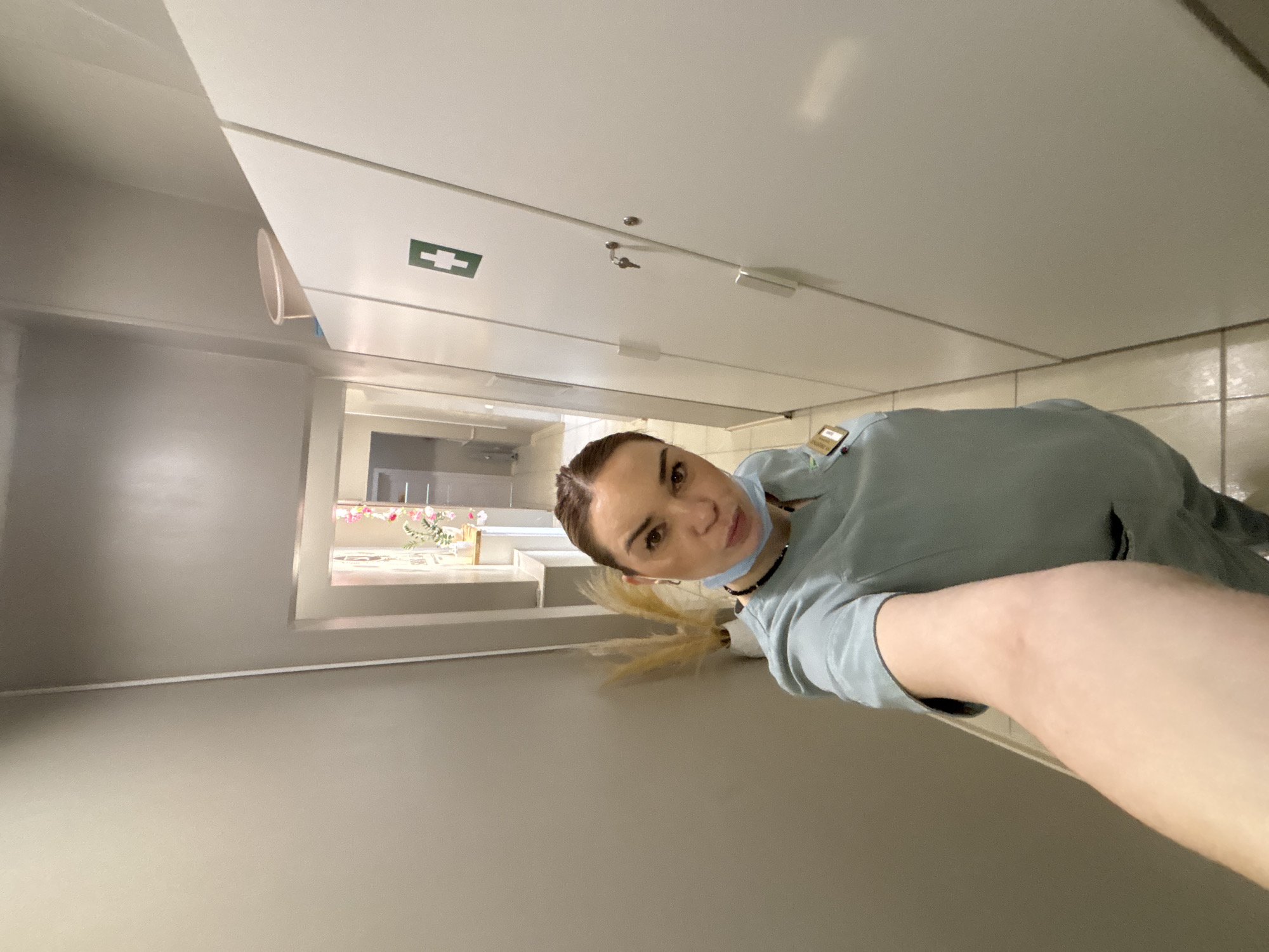 A low-angle shot depicts a woman wearing a blue top and a face mask, with her hair in a ponytail. She is looking at the camera. The background includes a white ceiling with a first aid sign and visible lighting fixtures. A window is visible, revealing an interior space with floral decorations and other decorative objects. The location is likely within a building in Vsevolozhsk, Russia.