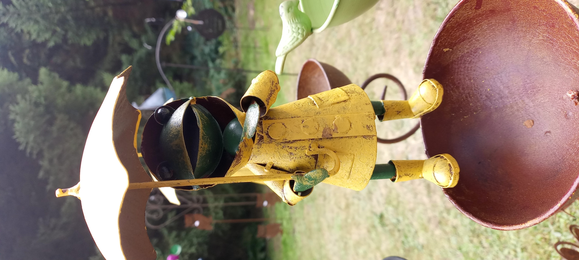 A metal garden decoration of a frog wearing a yellow raincoat and yellow boots, holding an open yellow umbrella, is perched on the rim of a rusty brown metal bowl. The frog has black eyes and an open mouth, revealing a green interior. In the background, out of focus, are green trees, other garden decorations, and a grassy area. The scene is an outdoor setting, possibly a garden.