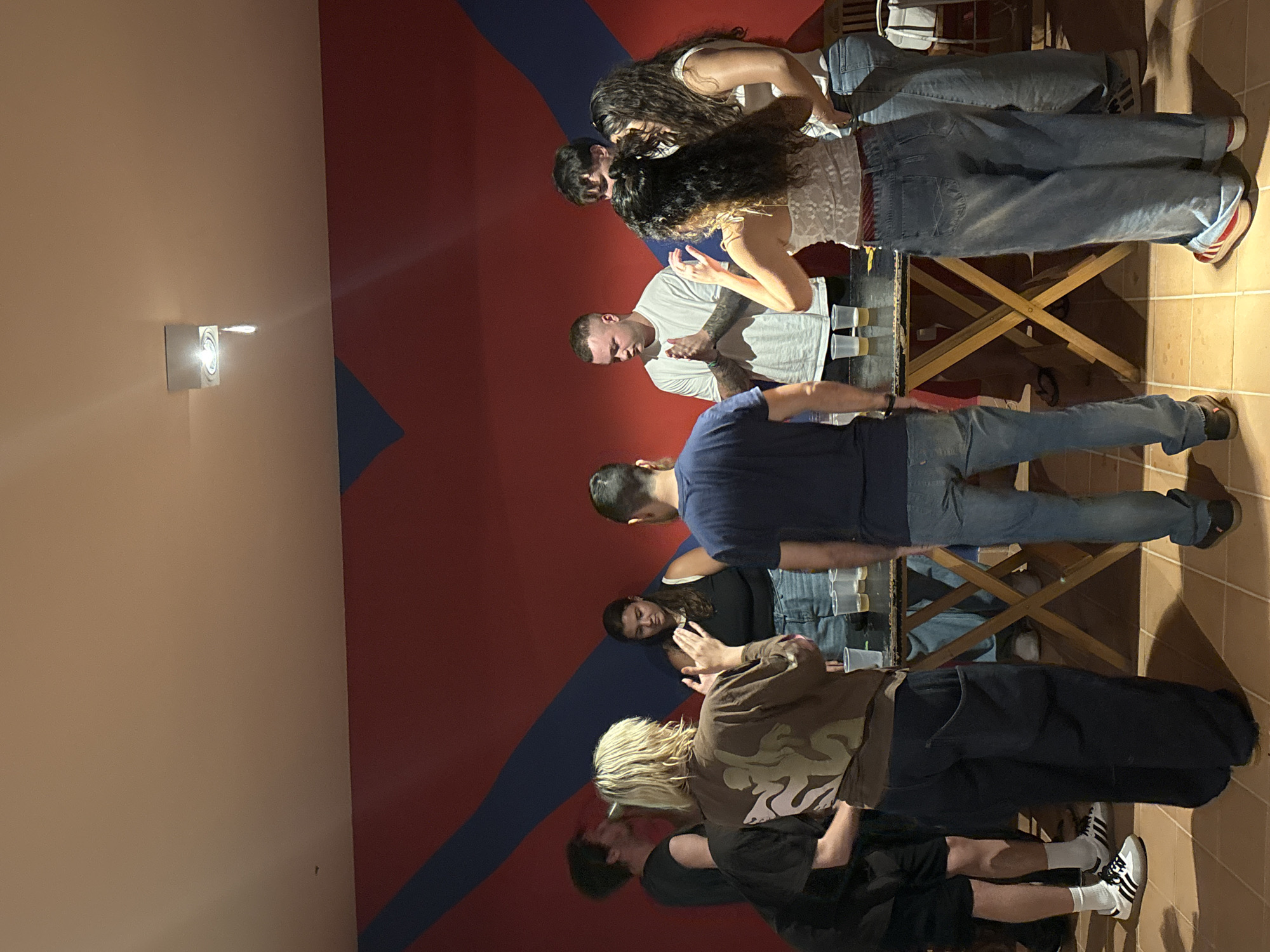 A group of people stands around a wooden table in an indoor setting. The walls are painted with a red and blue design. People are wearing casual clothing, including jeans, T-shirts, and sneakers. Some are facing the table, possibly interacting with objects on it, such as plastic cups. The floor appears to be tiled. A light fixture is visible on the wall. There is no evidence that the image was taken in São Paulo, Brazil.