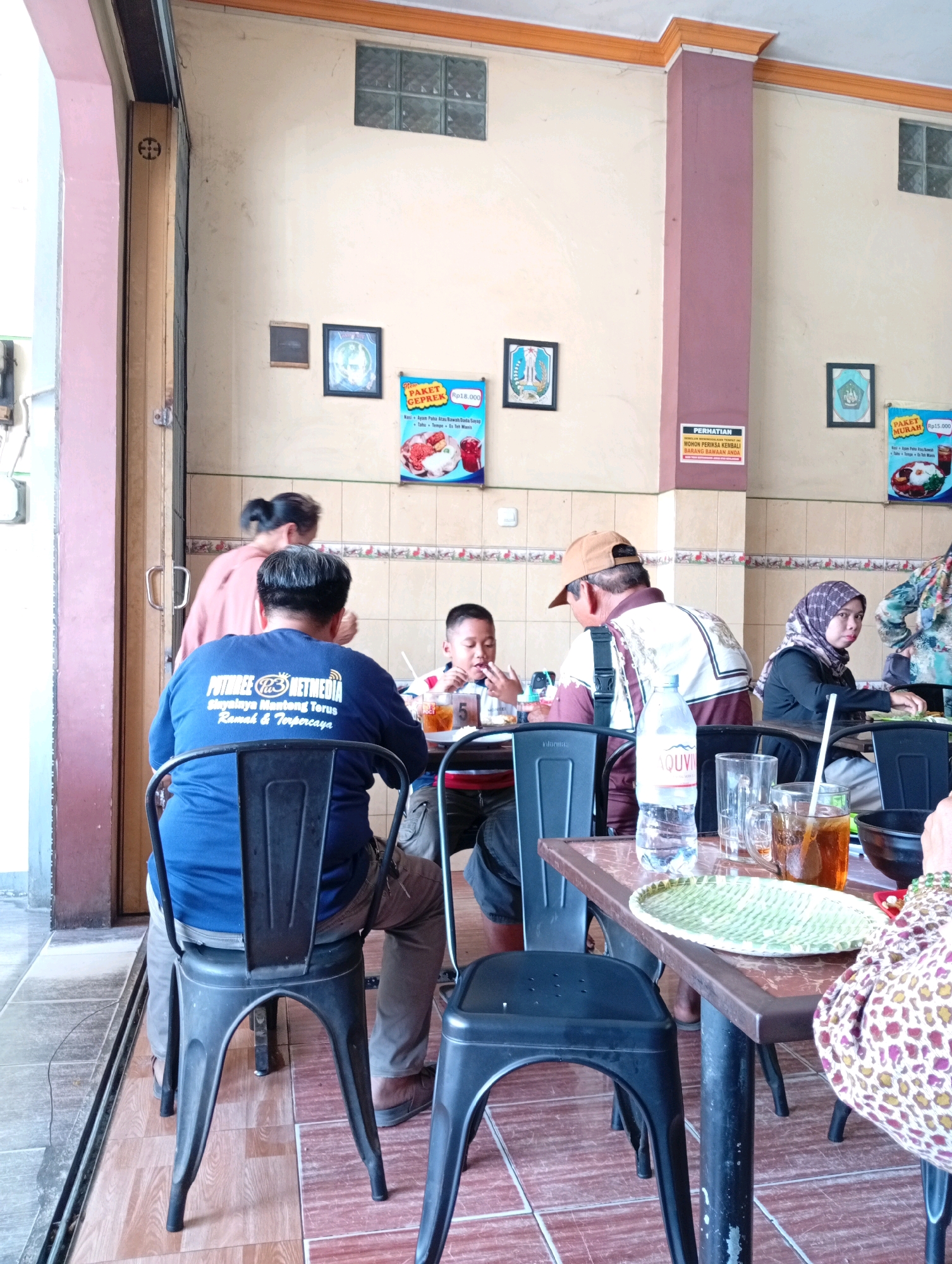 The image captures a scene inside a casual dining establishment, likely a restaurant or warung, in Kongsijaya, Indonesia. The environment appears to be indoors, with tiled flooring and a neutral-colored wall decorated with framed pictures and informational posters. Natural light enters from an open doorway on the left, illuminating the space.
Several individuals are present, seated at tables and engaged in dining. In the foreground, a person wearing a blue t-shirt with white lettering is seated at a black plastic chair, their back to the camera. To their right, a young boy with short dark hair is seated, facing forward and eating. A man in a brown baseball cap and a patterned shirt is seated behind the boy, also facing forward. Further to the right, a woman wearing a hijab and a patterned top is seated at another table, looking towards the camera. Another woman in a pink top is standing near the doorway, her back to the camera, appearing to be in motion.
On the tables, various objects related to dining are visible. There are plates with food, glasses of iced tea, a bottle of water, and possibly other beverages. The tables themselves appear to be made of wood with a patterned surface. The overall atmosphere suggests a casual and communal dining experience.