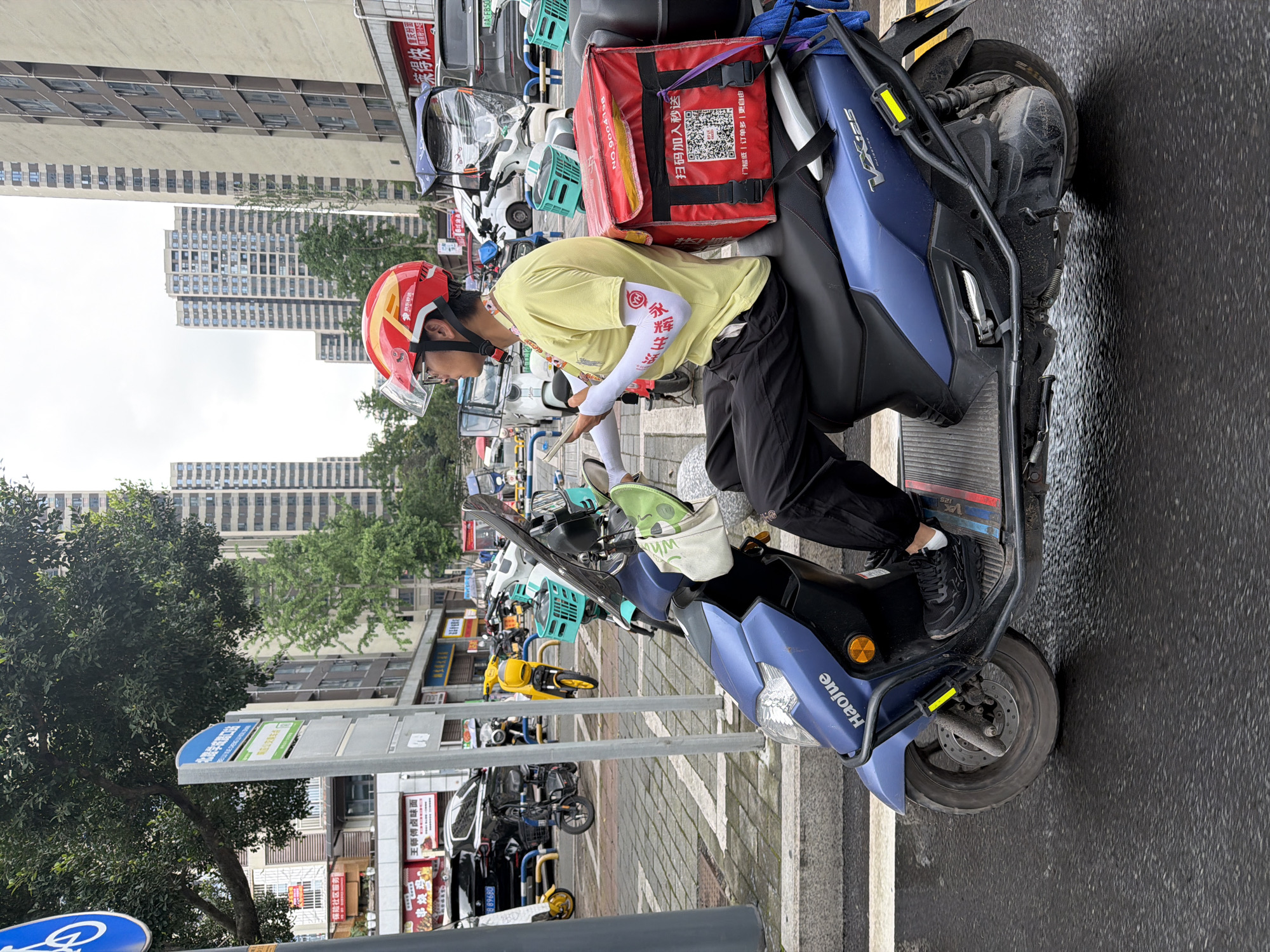 The image shows a delivery driver sitting on a blue Haojue VX125 scooter in Chongqing High-tech Zone, China. He is wearing a red helmet with a clear visor, a light yellow shirt, and black pants. White sleeves cover his forearms, with Chinese characters visible on them. He appears to be looking at a device in his hands, possibly a phone or delivery tablet.
The scooter has a red delivery bag attached behind the driver, with a QR code and Chinese characters printed on it. Various other scooters and vehicles are parked in the background, along with signs in Chinese. High-rise buildings can be seen in the distance, suggesting an urban environment. The sidewalk and road appear wet, indicating recent rain. A blue bicycle symbol is visible on a sign in the foreground. Street vendors and shops line the street.