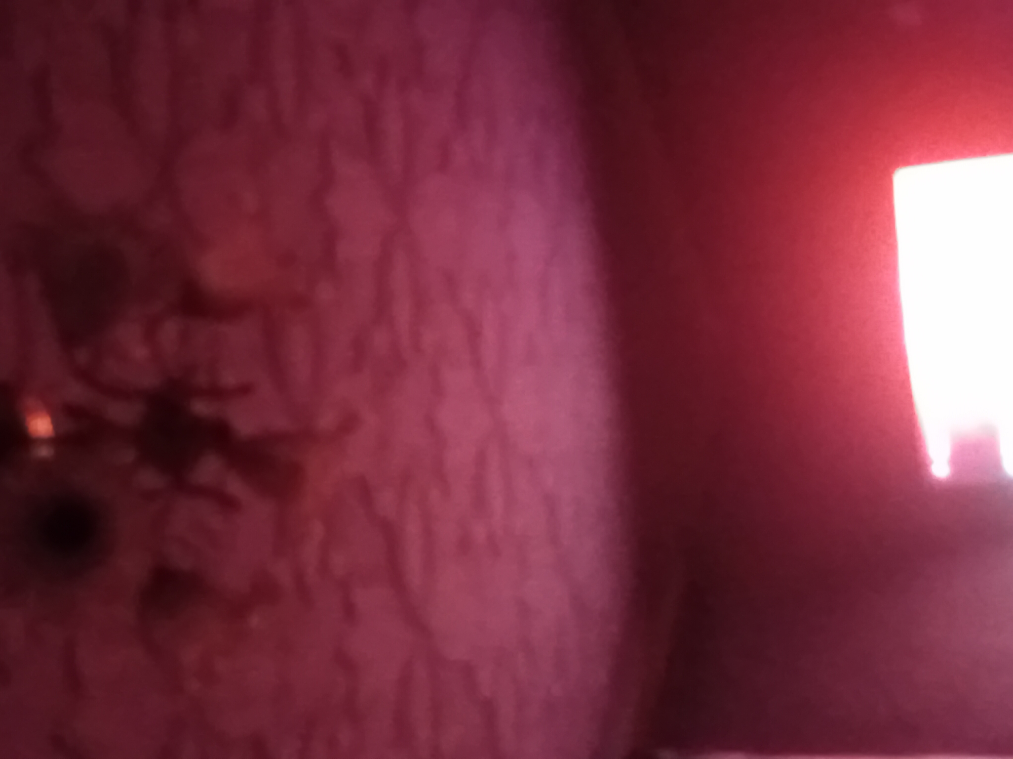 The image shows a dimly lit interior, likely a room, with a textured ceiling and a wall. The ceiling appears to have a floral or patterned wallpaper in shades of pink and red, with a dark, blurry object hanging from it, possibly a light fixture. The wall below the ceiling is a darker shade of red or maroon. In the lower right portion of the image, a bright light source is visible, casting a strong glow and creating a bright rectangle of light, suggesting a window or a light panel. The overall impression is of an indoor space with low lighting conditions. The image is blurry and lacks sharp details, making it difficult to identify specific objects or activities. There is no visible indication of the city of Verkhniachka, Ukraine.