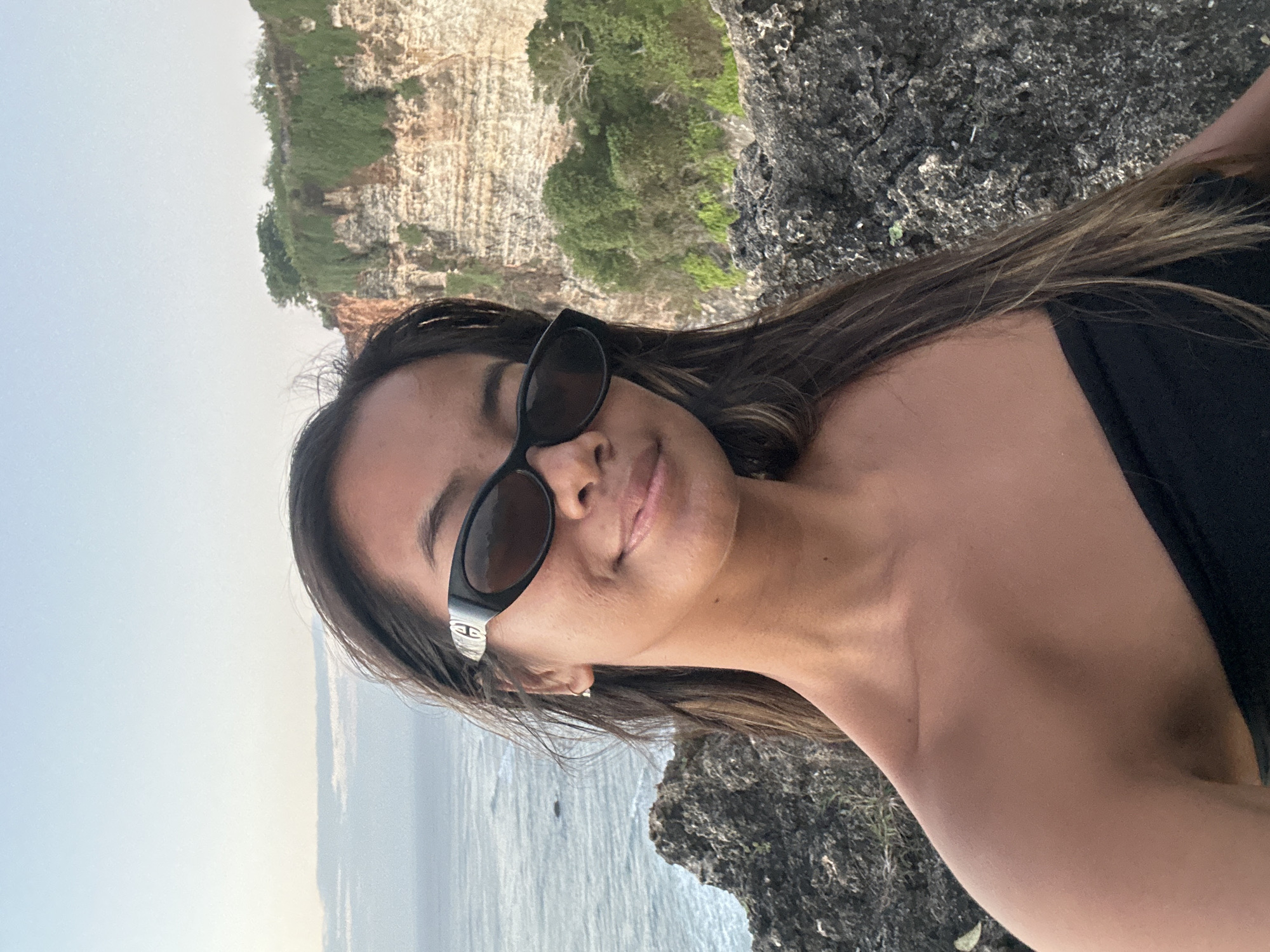 An outdoor, waist-up, medium shot shows a young woman with long brown hair and sunglasses posing for a selfie on a cliff overlooking the ocean. The woman is wearing a black strapless top and a pair of black oval-shaped sunglasses. Her right shoulder and upper arm are visible, along with part of her neck and chest. Behind her, a lush green cliff face rises, with trees dotting the top. The ocean, with gentle waves, extends to the horizon under a pale blue sky. Rocky terrain is visible in the foreground and to the right of the woman. The setting is likely a tourist destination near Kuta Selatan, Indonesia, given the landscape.