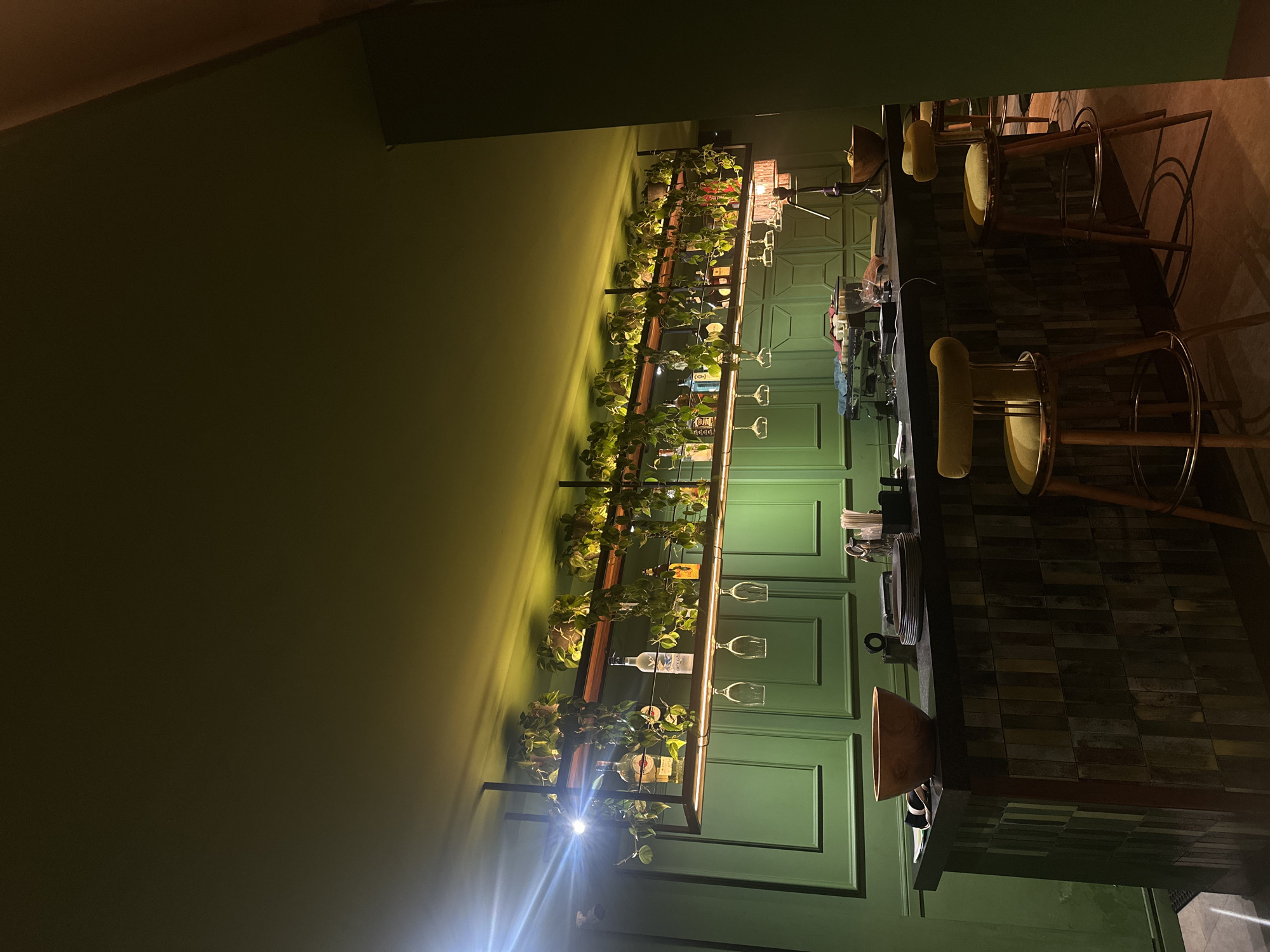 The image shows a bar interior in Ubud District, Indonesia. The walls are painted a deep green and decorated with a shelving unit filled with plants and bottles of alcohol. Wine glasses hang from the shelves. A dark bar runs along the wall, with yellow cushioned stools placed in front. The floor is tiled in a dark, geometric pattern. The scene is dimly lit, creating a relaxed, intimate atmosphere.