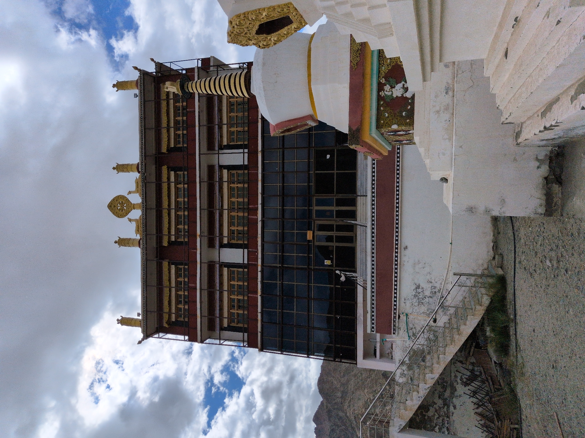 The image shows a view of the Phyang Monastery in Phyang, India, a Buddhist monastery located in the Indus Valley. The monastery is situated on a hillside and is characterized by its whitewashed walls, red and gold accents, and intricate architectural details. The main building of the monastery is a multi-story structure with numerous windows and decorative elements on its roof. In the foreground, there are several stupas, which are Buddhist reliquary monuments. One prominent stupa in the foreground is white with a striped conical top and colorful decorative bands. Adjacent to the stupa, there are stairs leading up to the monastery. The sky is cloudy with patches of blue, suggesting it may be a partly sunny day. The surrounding landscape appears to be mountainous and arid, typical of the region.