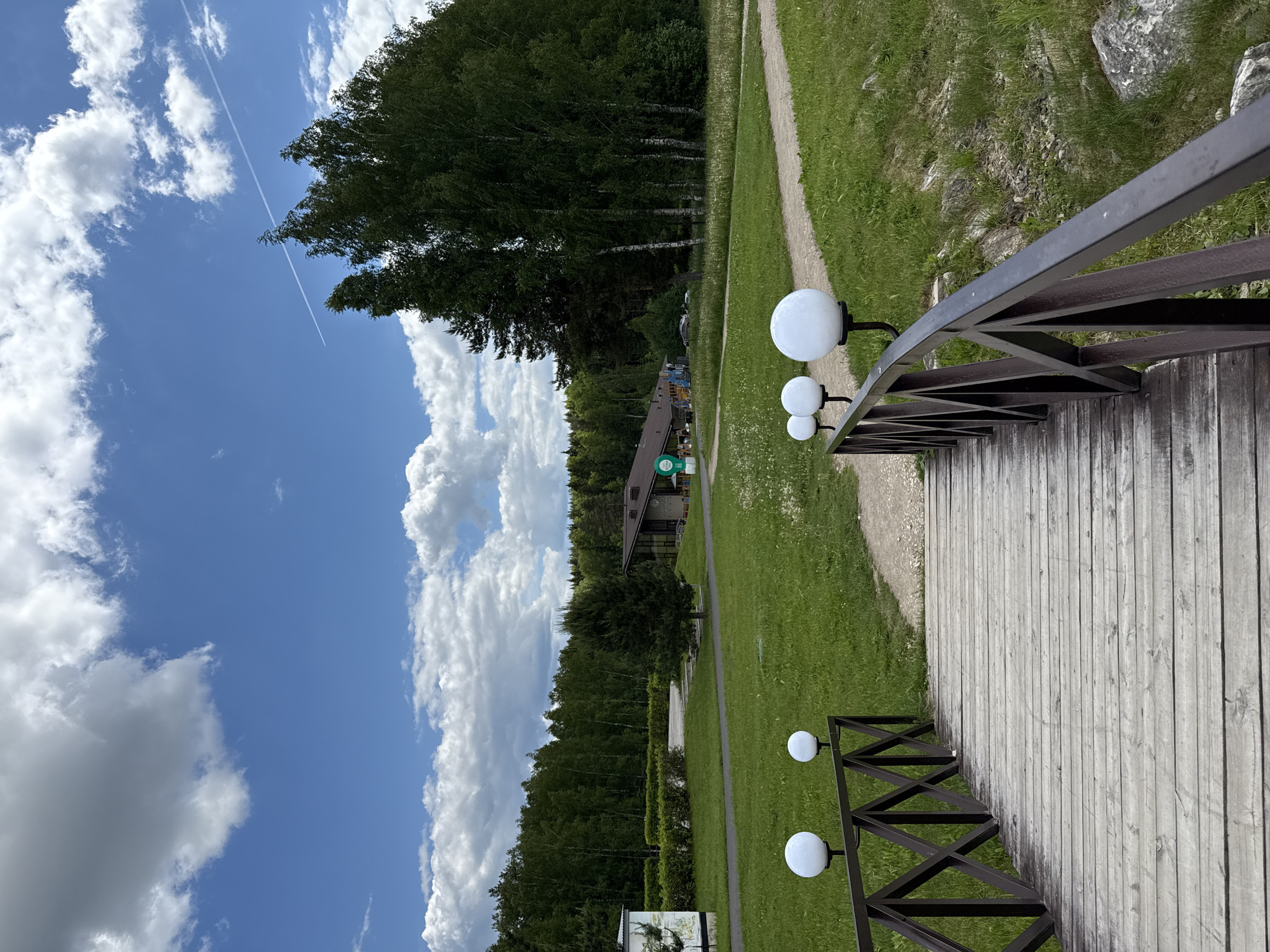 The image depicts a serene outdoor scene, likely within a park or resort setting in Dobrograd, Russia, captured on a bright, sunny day. The camera is oriented to show a wide view, with a clear blue sky dominating the upper portion and a natural landscape filling the rest of the frame.
In the foreground and extending into the midground on the right, there is a light-colored wooden walkway or bridge. A dark brown metal railing with geometric patterns lines one side of this structure. Several round, white globe-shaped lamps are attached to the railing at intervals, providing illumination. To the left of the walkway and along its edges, there are vibrant green grassy areas, interspersed with a dirt or gravel path that parallels the wooden structure.
In the midground and background, a dense line of lush green trees, including tall deciduous trees like birches, forms a natural backdrop. Nestled amongst these trees, a building with a dark-pitched roof and large windows is visible, suggesting a facility within the complex. A small green sign, possibly a logo or directional sign, is affixed to this building, though the text is not legible. Another small, unreadable sign is partially visible on the ground in the bottom left corner.
The sky is a brilliant blue, adorned with scattered, fluffy white cumulus clouds. A long, thin white contrail, indicating the passage of an aircraft, stretches across the upper part of the sky from left to right. The overall impression is one of peaceful natural beauty and well-maintained grounds, with no direct human activity visible. The bright lighting indicates it is daytime, likely midday or early afternoon, under clear weather conditions.