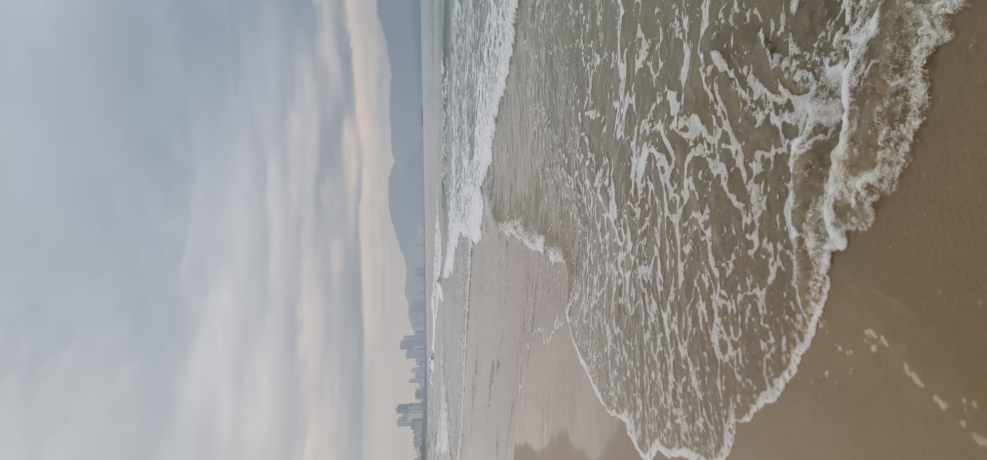 The image depicts a beach scene in Đà Nẵng, Vietnam. The foreground shows the shoreline where waves are washing onto the sand, creating foam. In the distance, a coastline with buildings is visible, partially obscured by haze. Behind the buildings, there are mountains. The sky is overcast with thick clouds. There are no people visible in the frame.