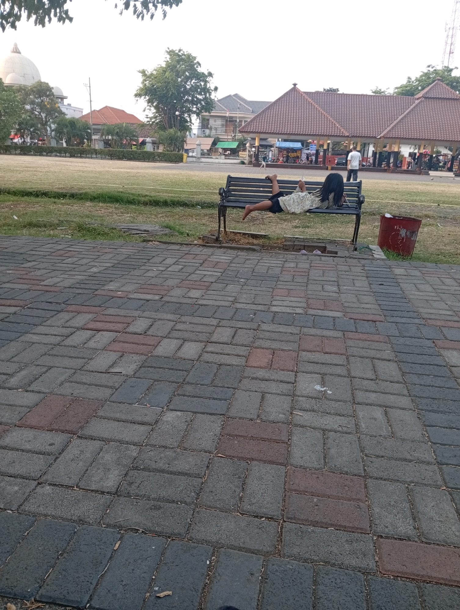 In the city of Bangil, Indonesia, a young child with dark hair lies on their back on a park bench. The child is wearing a light-colored, patterned shirt and dark shorts. To the right of the bench, a red trash can is visible. The park features a paved walkway in the foreground, made of interlocking bricks in shades of gray and rust. Beyond the walkway and bench, a grassy field is present, bordered by a line of manicured bushes. In the background, several buildings with terracotta roofs are visible, along with trees and other park elements. A person in a white shirt stands in the middle ground, facing away from the camera. The sky is bright and overcast.