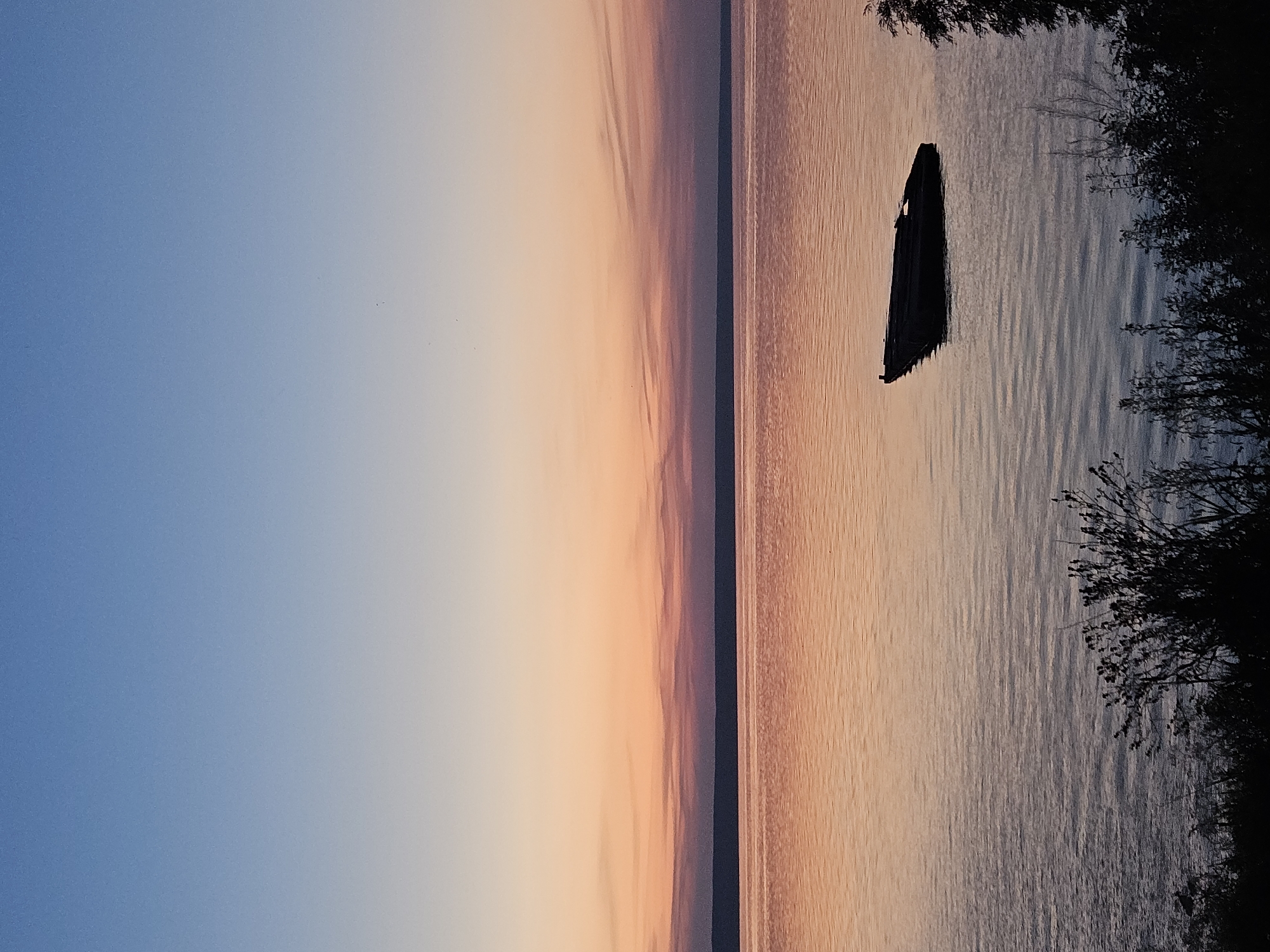 A long, dark, dilapidated boat is submerged in calm water during a sunset. The sky above transitions from a deep blue to a pale orange, with hints of pink and purple. The water reflects the colors of the sky, creating a warm, golden hue. Silhouetted trees line the bottom of the frame. The scene suggests a quiet, serene evening in a natural setting. The location of Leninsky, Russia, is not depicted or suggested by any visual cues in the image.