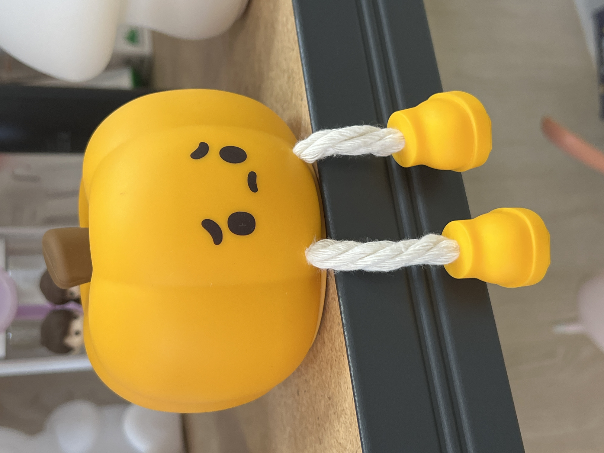 A close-up, eye-level shot shows a bright orange, plump pumpkin figurine with a sad face made of black ovals and a curved line. The figurine is sitting on a black shelf that has a textured surface. Two white braided cords hang down from the bottom of the pumpkin, each ending in a small, yellow, bell-shaped foot. The background is blurred, with hints of other decorative items, including what appears to be a white mushroom-shaped object and small figurines with brown hair. The lighting is soft and even, highlighting the smooth texture of the pumpkin. The scene is an indoor setting, likely a shelf in a home or shop. There is no discernible connection to Ridder, Kazakhstan.