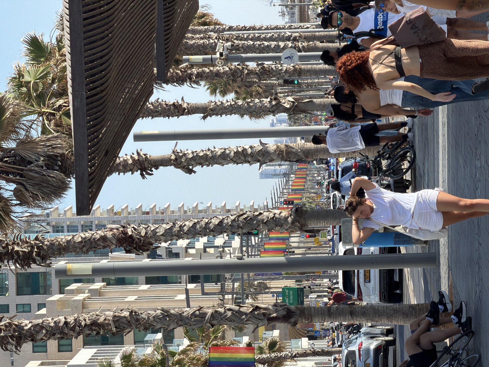This image depicts a bustling street scene in Tel-Aviv, Israel, specifically along a promenade or major thoroughfare near the coast. The environment is characterized by a clear blue sky and abundant sunlight.
In the immediate foreground, a young woman with light brown hair stands, facing slightly left, with her hands raised to adjust her hair. She wears a white t-shirt, white shorts, and carries a white shoulder bag. To her right, another woman with curly red hair is seen from behind, walking away from the camera. She wears a black strappy top and leopard-print pants, carrying a large brown tote bag with partially visible text "SCHAF". Behind her, a woman in a white strapless top and blue jeans walks in the same direction. Further right and slightly behind, a man with sunglasses and a white Reebok tank top walks towards the camera, carrying a white backpack. On the far left foreground, the legs of a person seated on a bench are visible, wearing black shorts and black sneakers; a bicycle leans nearby.
The street is lined with numerous tall palm trees on both sides, and a series of modern, multi-story buildings with balconies and glass windows rise in the background. Interspersed among the palm trees are tall, grey street poles, some fitted with light fixtures, others with surveillance cameras and speakers. A modern wooden pergola structure with curved slats extends over the sidewalk on the right side, providing shade.
Vehicular traffic is present on the paved road, which features multiple lanes. A silver sedan is partially visible on the left, and a white SUV with an Israeli license plate "572-44-647" is visible further back on the right. Numerous rainbow flags, indicative of LGBTQ+ pride, are prominently displayed along the street, hanging from poles and strung overhead, extending into the distance on the left side. A green street sign with Hebrew text is visible on the left, along with a blue circular sign on a pole depicting a bicycle, indicating a cycling path. Traffic lights, both red and green, are visible in the distance. Many other pedestrians populate the sidewalks and crosswalks, indicating a lively urban area.
