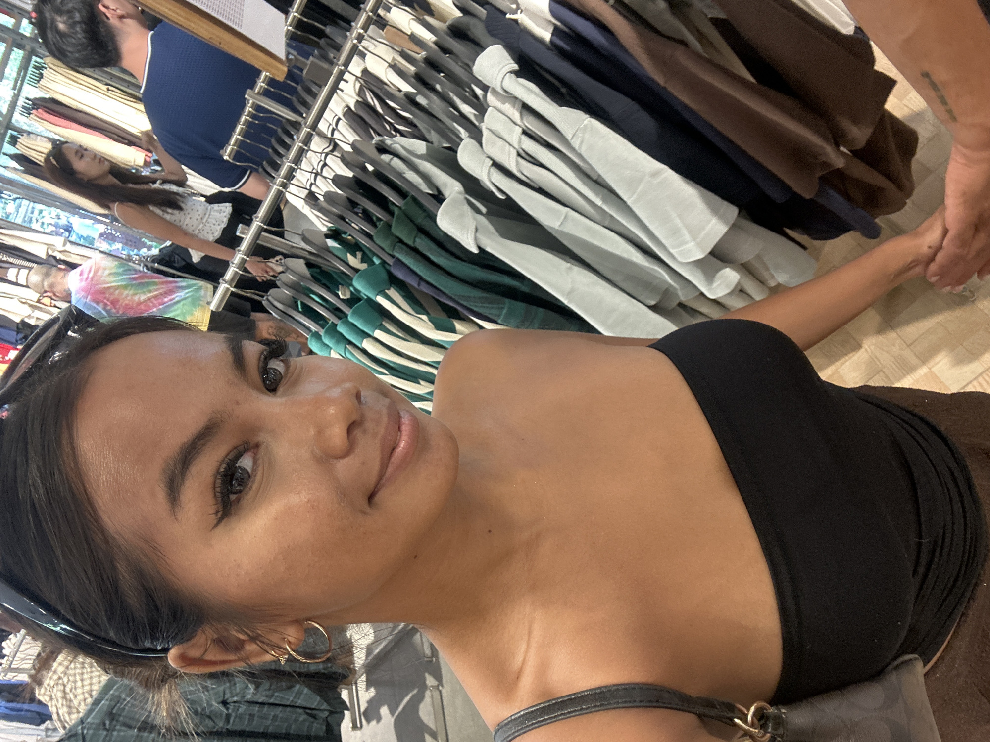 A young woman with dark hair pulled back, wearing a black strapless top and gold hoop earrings, is captured in a selfie. She has distinctive winged eyeliner and a light mole on her collarbone. Her right arm is visible, holding a small gray purse. In the background, clothing racks display various garments, suggesting a retail environment, possibly a clothing store. The lighting appears to be indoor, with some natural light filtering in. The overall scene suggests a casual shopping outing in Kuta, Indonesia.