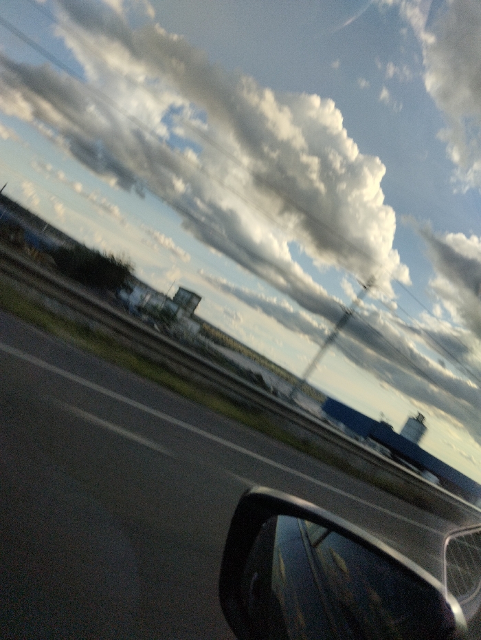 The image shows a view from the inside of a car driving on a highway in Athi River, Kenya (latitude: -1.495, longitude: 37.063). The car is moving fast, as indicated by the blurred view of the surroundings. The sky is mostly clear, with some white cumulus clouds floating across the blue background. To the right of the highway, a blue building, likely a factory or warehouse, can be seen. The surrounding area appears to be industrial or commercial, with visible vegetation on the side of the road. The scene is a typical urban landscape of a developing country, characterized by fast-paced development and busy roads. The view from the inside of the car suggests a moment of casual observation and a sense of movement through the cityscape.