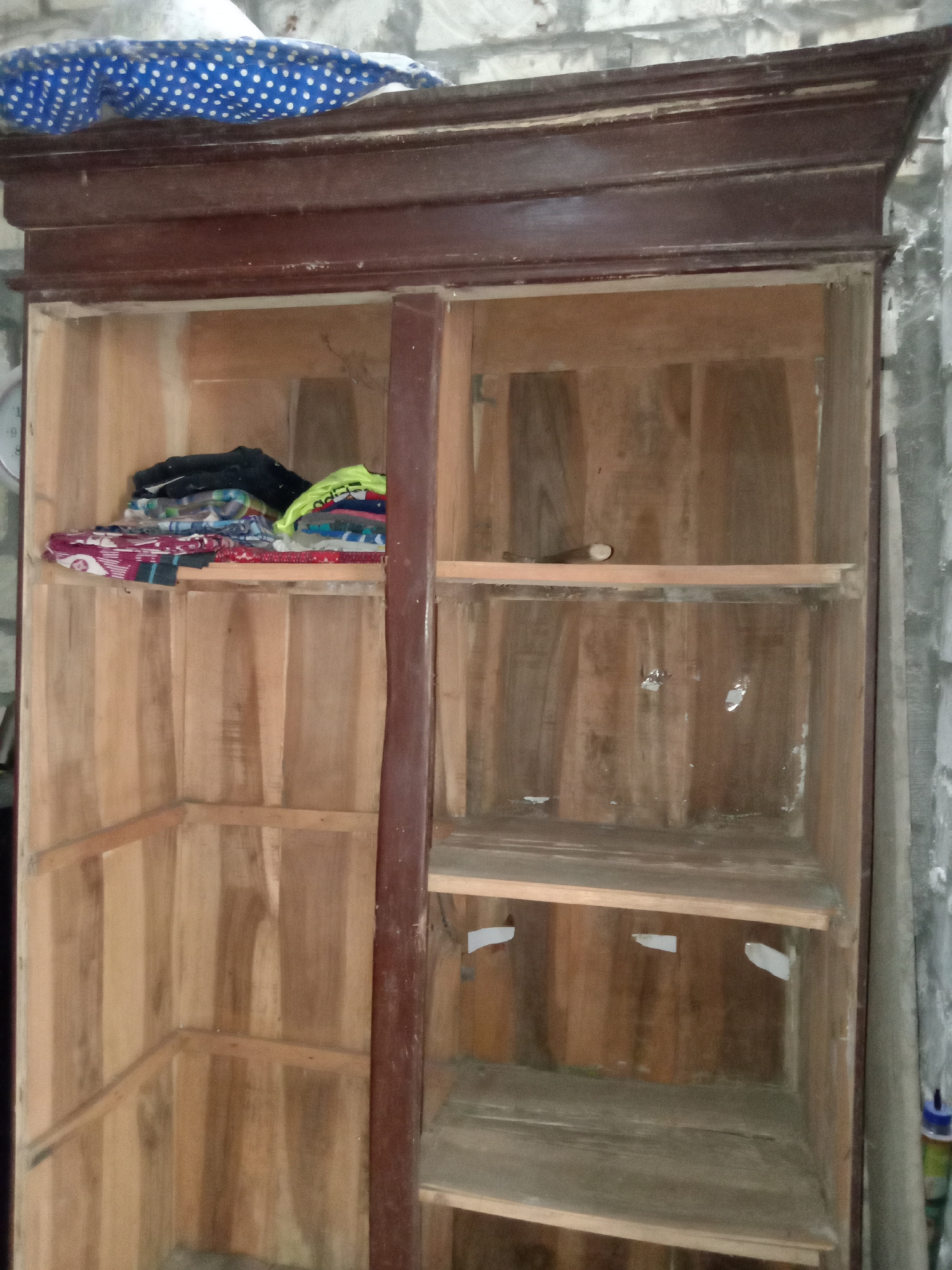 An indoor, eye-level, medium shot captures an open wooden wardrobe. The wardrobe has a dark brown frame with a decorated top edge. Inside, the wood is a lighter, natural tone. The left side of the wardrobe is empty except for a pile of folded clothing on the top shelf, which includes items in black, white, blue, red, and green patterns. The right side of the wardrobe has three empty shelves. A blue and white polka dot fabric item rests on top of the wardrobe. The background consists of a rough, grey concrete wall. The scene is located in Jumput, Indonesia.