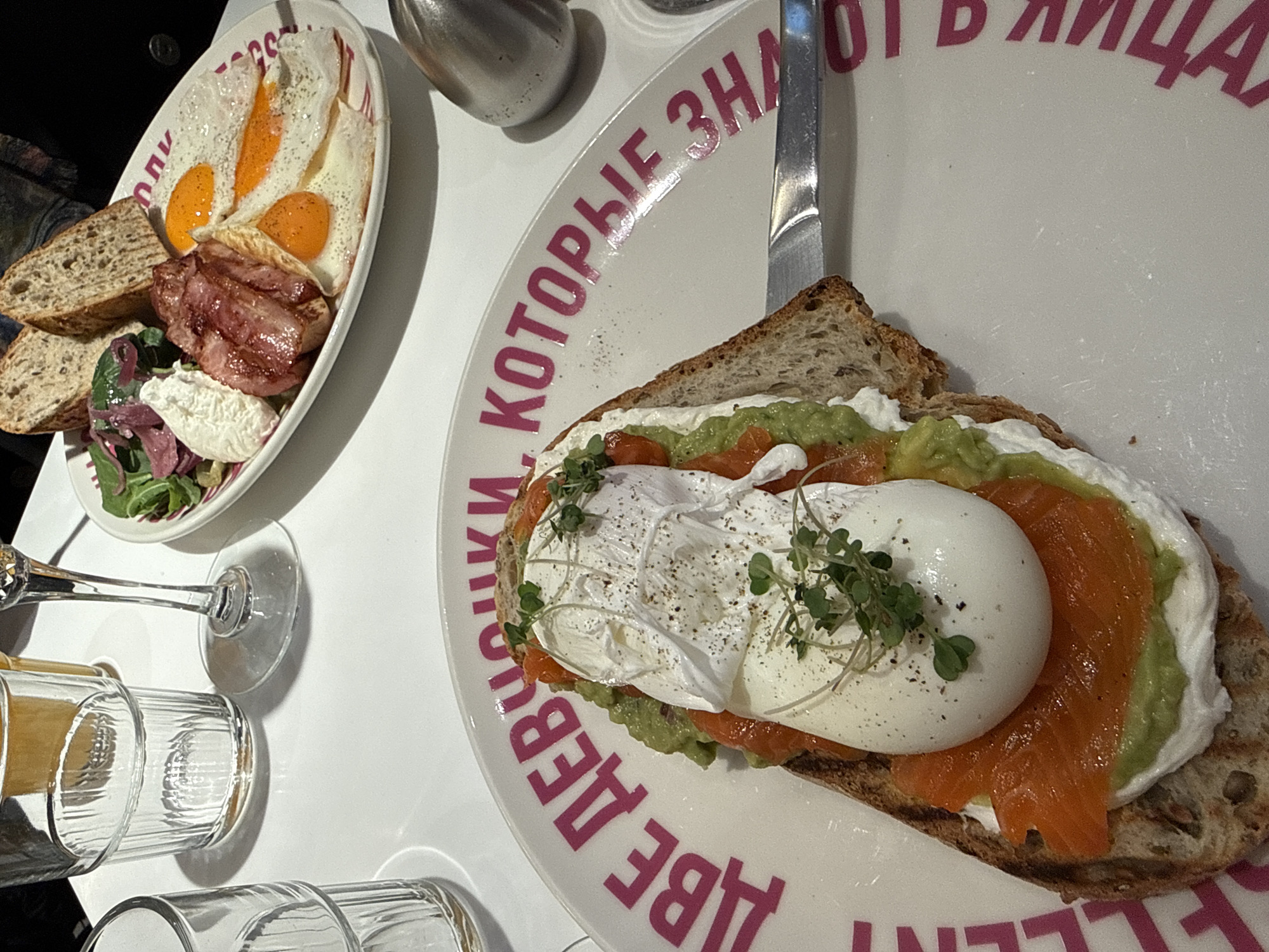 The image shows two plates of food on a white table, likely in a restaurant in Moscow, Russia, indicated by the text in Russian on the plates. The plate on the left contains fried eggs, bacon, and bread, with a small salad. A silver container and glassware are also present. The plate on the right has a slice of bread topped with avocado, salmon, and a poached egg, garnished with greens. A silver knife rests beside it. The scene appears to be a close-up shot of a breakfast or brunch setting.