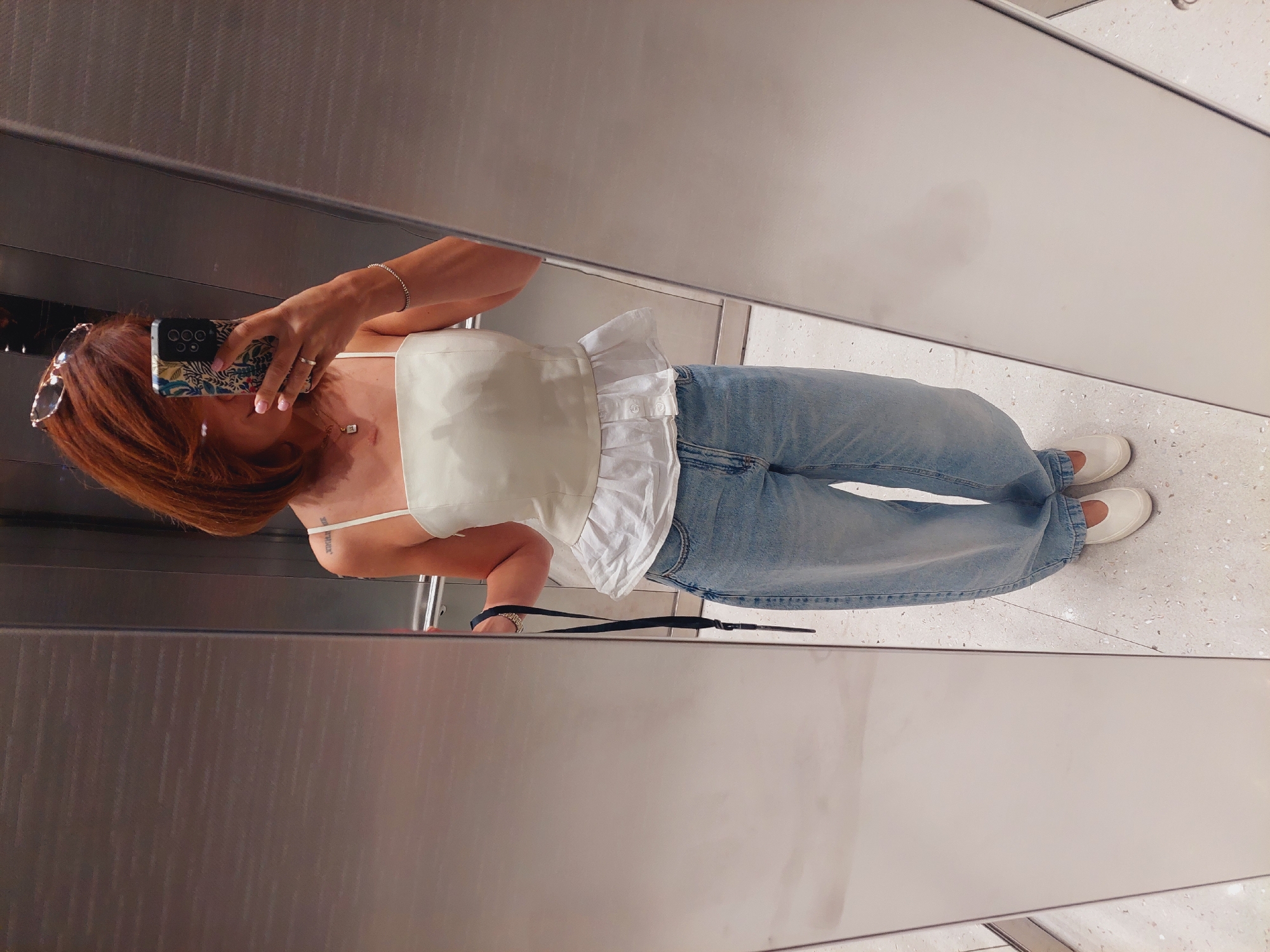 A woman with reddish-brown hair is standing in front of a mirror in what appears to be an elevator. She is wearing a white peplum top with spaghetti straps and light blue, wide-leg jeans. She is holding a dark strap in her right hand and a mobile phone in her left hand, taking a selfie. She has a necklace and bracelet on, and sunglasses are perched on her head. The elevator walls are metallic and reflective. The floor is speckled light gray and white. This scene is not identifiable as being in Sofia, Bulgaria, or any other specific city.