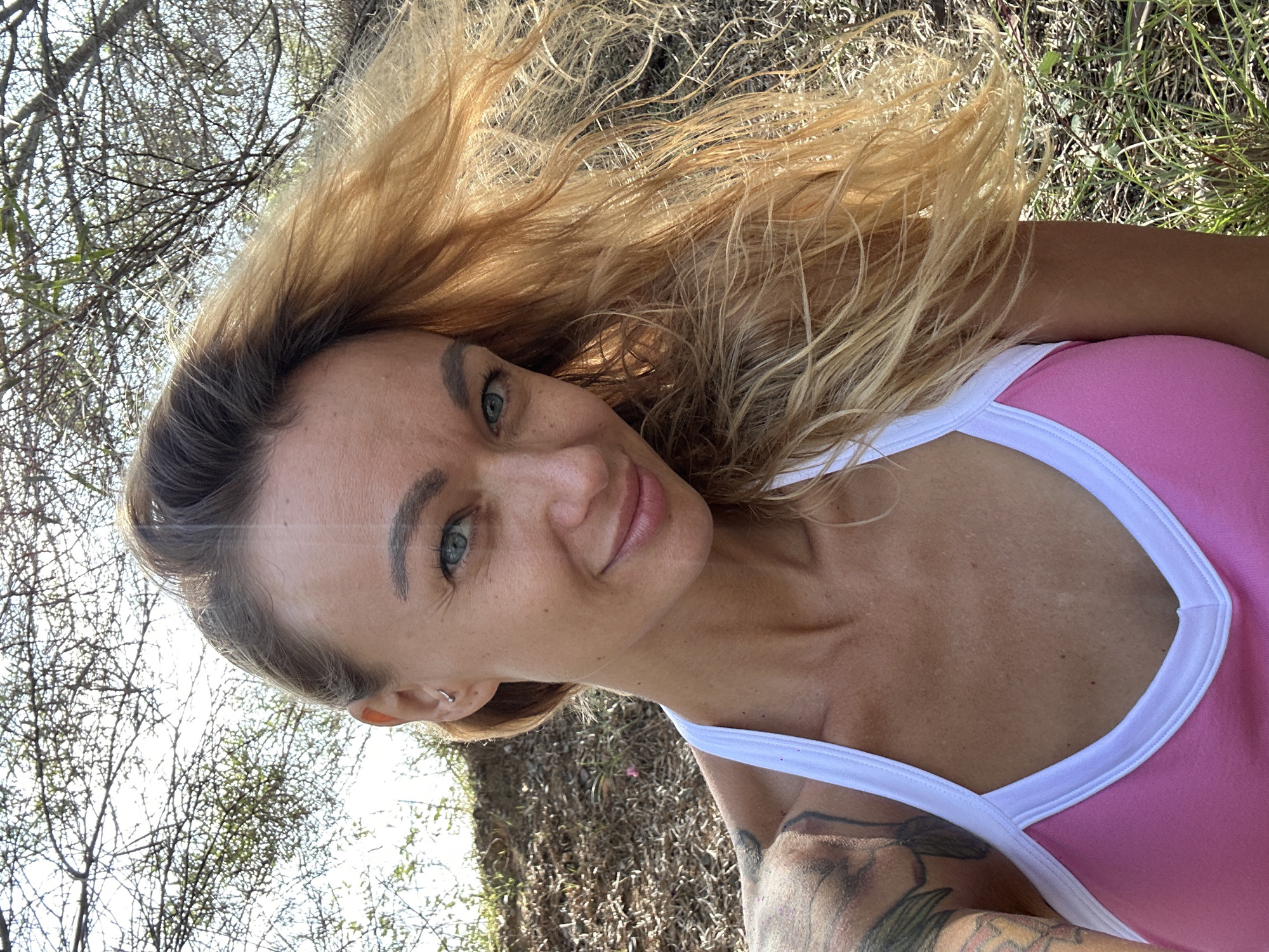 A close-up, low-angle, selfie-style shot shows a fair-skinned woman with long, wavy blonde hair against a backdrop of dry vegetation and sparse trees. The woman, who appears to be in her 30s, has striking blue eyes and a subtle smile. She is wearing a pink tank top with white trim. A tattoo is visible on her left shoulder and upper arm. The image is taken outdoors, likely in a rural or natural setting, as indicated by the dry, uncultivated ground, grass, and trees. The bright sunlight creates lens flare and highlights the woman's hair and skin. The location in Phu Thuy Ward, Vietnam, is not discernable from the visual information provided in the image itself.