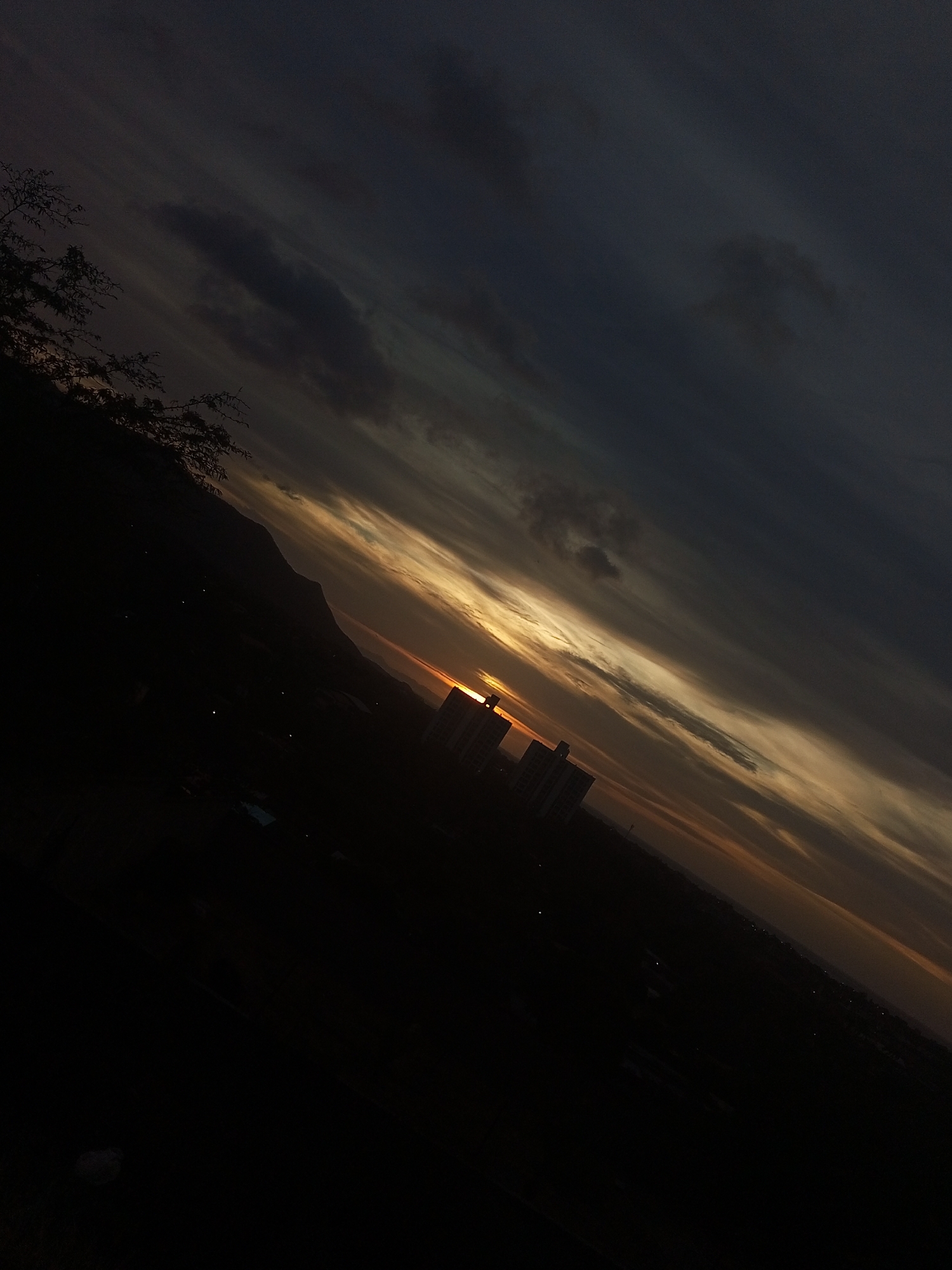 The scene depicts a tilted outdoor view of a sunset or sunrise over Juan Griego, Venezuela. The sky occupies the upper two-thirds of the frame, characterized by stratified clouds transitioning from dark grey and blue-black in the upper right to streaks of orange, yellow, and lighter grey along the horizon. The sun is partially visible as a bright orange-yellow orb, positioned low on the horizon, bisected by the upper portions of two silhouetted, multi-story buildings. A dark, silhouetted tree branch with sparse foliage is present in the upper-left quadrant. To the left of the prominent buildings, a large, dark, mountainous landmass is silhouetted against the lighter sky. The lower portion of the image, below the mountain and buildings, is predominantly dark, with scattered small points of light indicating urban illumination. No individuals or discernible actions are present.