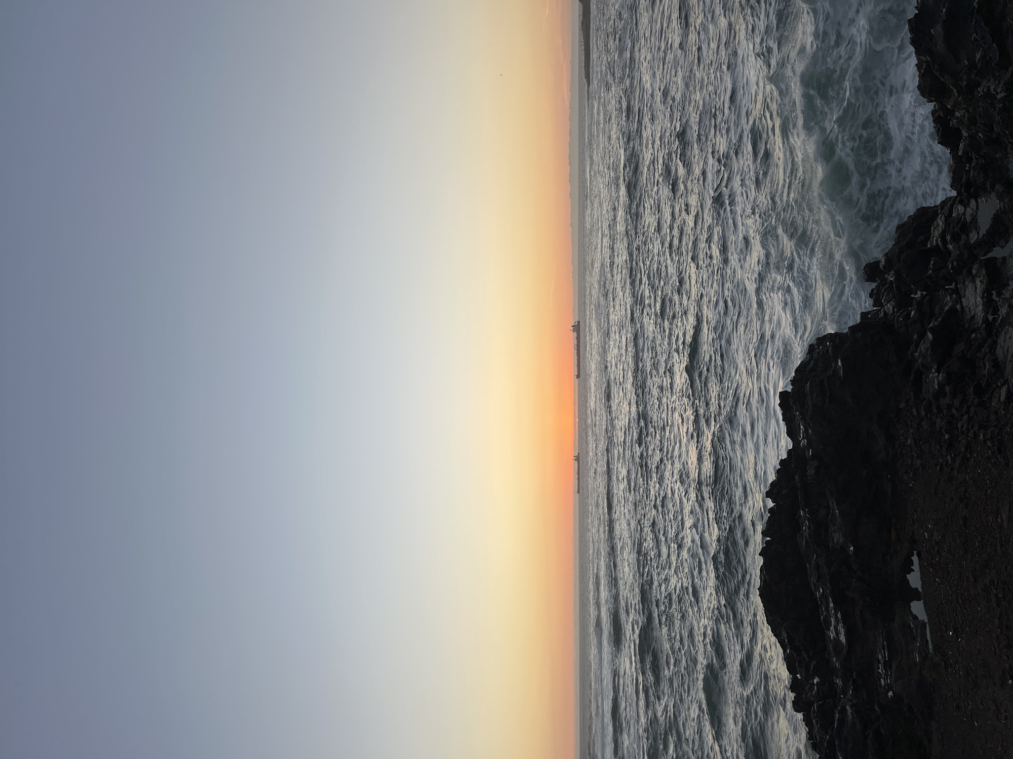 The image depicts a scene at the coastline. The sky transitions from light blue to a vibrant orange sunset. The ocean is churning with waves, bordering a rocky area. Arica, Chile, is discernible. There are ships on the horizon.