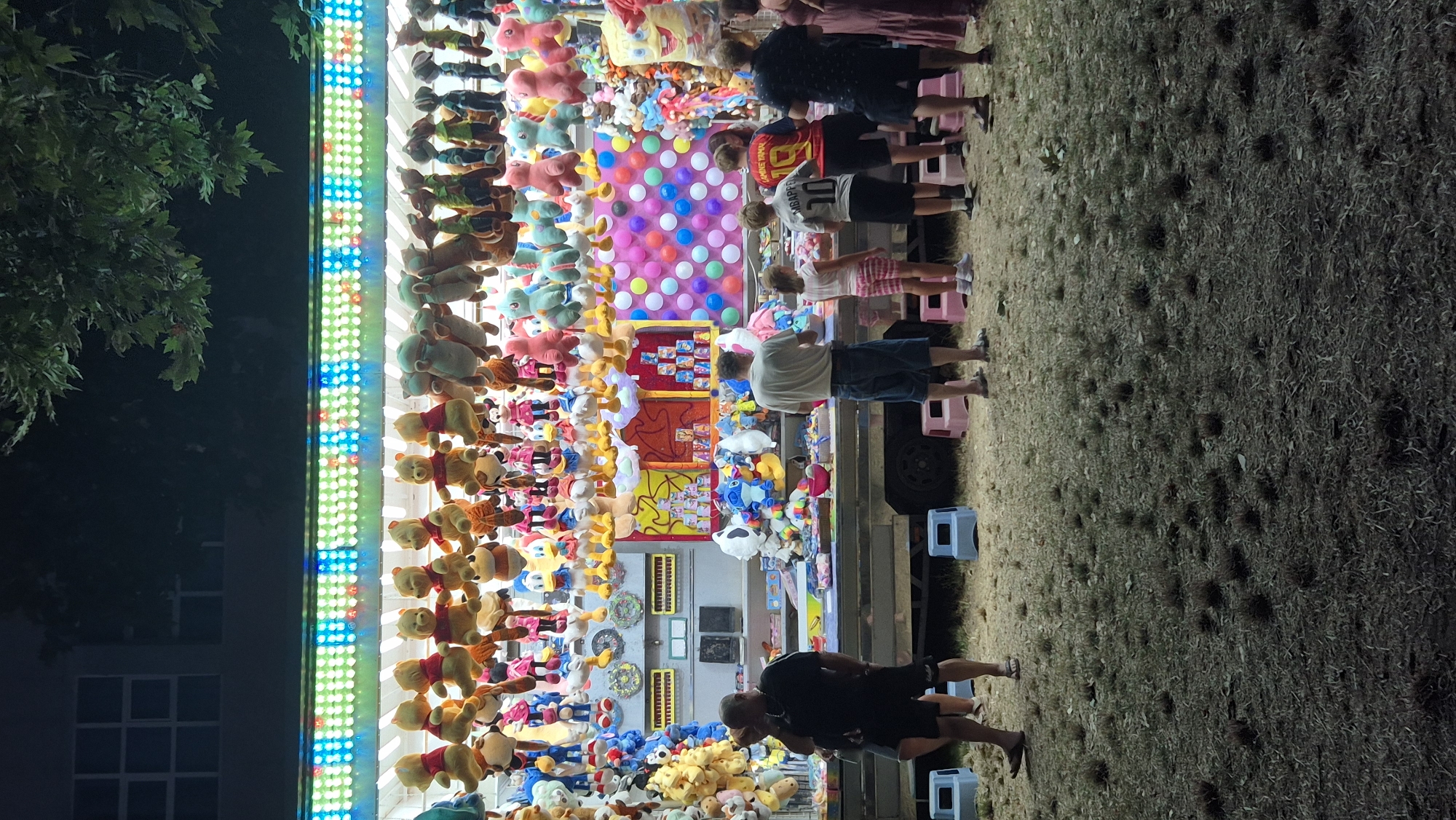 This image depicts a fairground stall at night, likely located in Primorsko, Bulgaria, based on the context provided. The stall is brightly lit with rows of colorful LED lights along its top edge. It is heavily stocked with an assortment of plush toys, predominantly Winnie the Pooh characters and other cartoon figures, hanging from above and displayed on shelves. Several people are standing in front of the stall, observing the merchandise or possibly waiting to play a game. The ground in front of the stall is covered with dry grass and scattered debris. To the left, a building with a window is visible. The overall scene suggests a festive atmosphere common at fairs or carnivals.