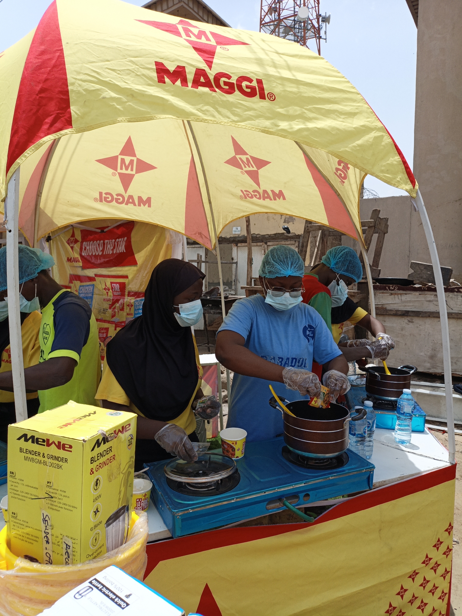 This media file captures an outdoor promotional event for Maggi products in Tarauni, Nigeria. The scene is set under a large yellow and red Maggi-branded tent. Several individuals, likely employees or volunteers, are actively participating. Two people in the foreground, wearing blue hairnets and face masks, are preparing food. One person, wearing a blue t-shirt with "PL PARADOL" printed on it, is adding a packet of Maggi seasoning into a pot on a portable gas stove. Another person, partially visible in a yellow shirt and black hijab, is also wearing a face mask and seems to be assisting with the cooking. To the left, another individual in a yellow shirt and blue hairnet is also wearing a face mask.
Several objects are prominent in the scene. To the left, a yellow box labeled "MeWe Blender & Grinder" is visible, along with other Maggi product packaging in the background. Water bottles, cups, and cooking utensils are arranged on a white table in front of the food preparation area. A blue gas stove is the central focus of the cooking activity. The overall atmosphere appears to be a promotional setup, possibly a cooking demonstration or sampling event. The bright sunlight suggests it is daytime, and the presence of the tent indicates an outdoor event. The background shows a slightly cluttered environment with some construction materials, suggesting a less developed area or a street-side location. The text visible includes "MAGGI", "CHOOSE THE STAR", "MeWe Blender & Grinder", and various product specifications in English, French, and Portuguese.