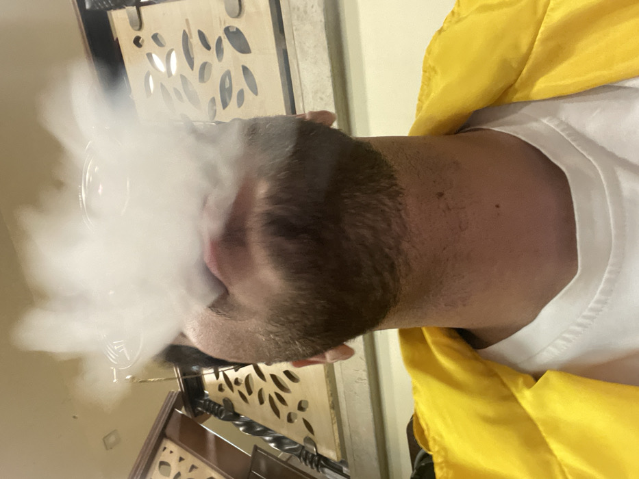 The image displays an upward-angled view of an adult male within an indoor establishment, identified as being in Homs, Syria. The subject, who possesses a trimmed beard on his chin and jawline, is observed exhaling a substantial volume of dense, white vapor from his mouth, which largely obscures his upper facial features. A clear, rounded object, consistent with a shisha mouthpiece, is partially discernible within the vapor plume directly in front of his mouth. He is attired in a white crew-neck t-shirt underneath a bright yellow collared jacket. The background reveals interior architectural elements: a light-colored wall on the right featuring a decorative wooden panel with multiple irregular, elongated cut-out patterns, and a darker wooden structure on the left with turned vertical elements and carved details, indicative of a partition or railing. A square recessed light fixture is visible on the light-colored ceiling. The overall environment suggests a cafe or shisha lounge.