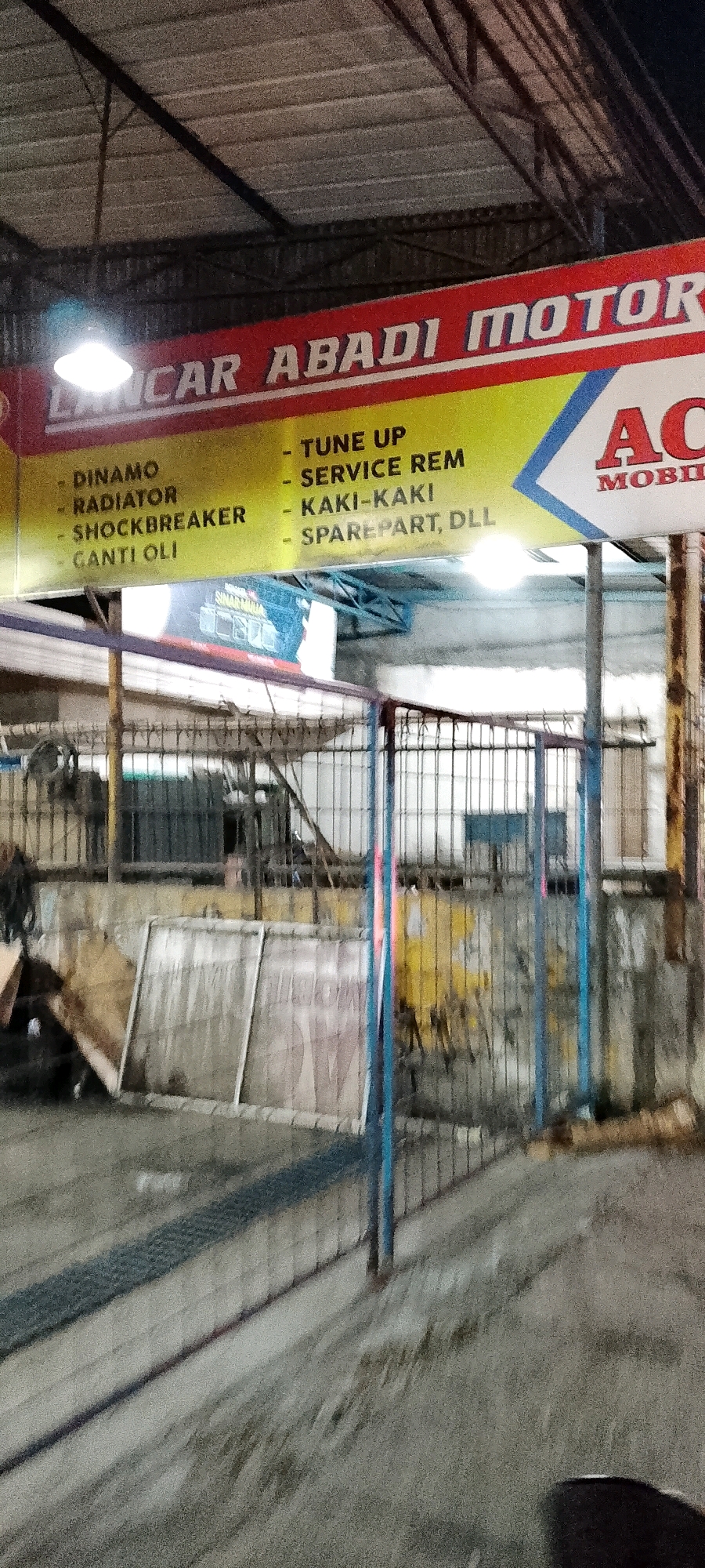 The image captures a storefront for a business identified as "Lancar Abadi Motor" in Gunung Putri, Indonesia. The storefront features a prominent yellow sign with red lettering at the top, displaying the business name. Below the name, a list of services is presented in black text on the yellow background. These services include "DINAMO," "RADIATOR," "SHOCKBREAKER," "GANTI OLI," "TUNE UP," "SERVICE REM," "KAKI-KAKI," and "SPAREPART, DLL." To the right of the list of services, the word "ACE" in large red letters with "MOBIL" in smaller red letters below it is visible, suggesting a connection to automotive parts or services. The overall scene depicts an outdoor, possibly nighttime setting with artificial lighting illuminating the sign. The background shows a fenced area with various materials and structures, suggesting a workshop or storage area behind the storefront.