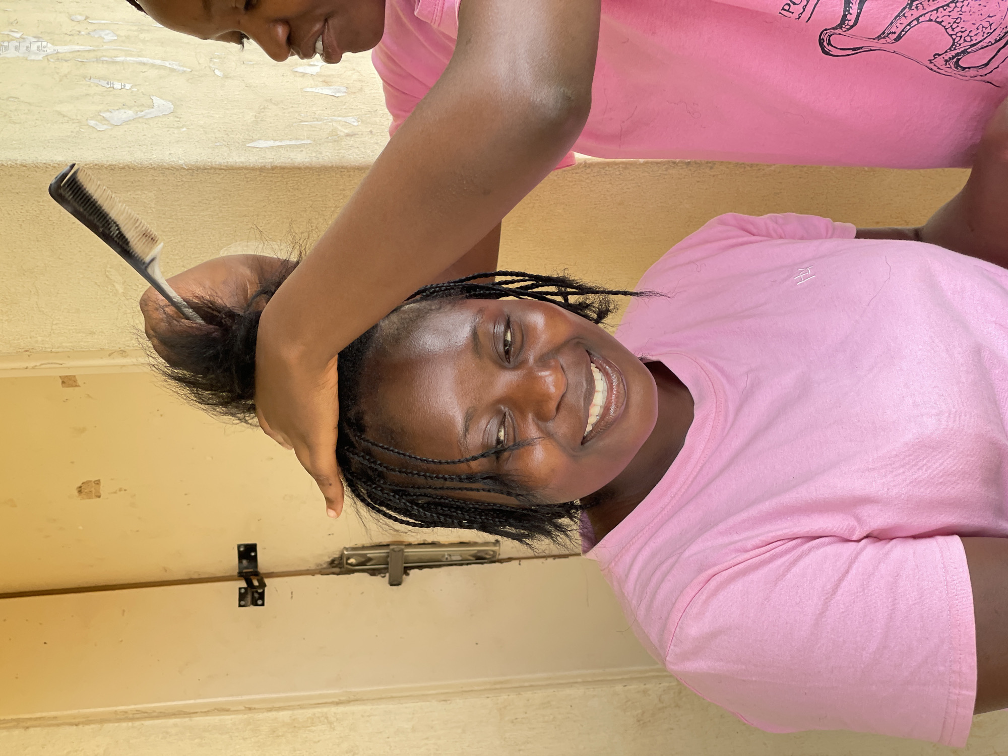 In Durumi, Nigeria, two young Black women, both wearing bright pink t-shirts, are the subjects of this vertical, outdoor photograph. The woman in the foreground, with her head tilted back and a wide smile, is having her hair styled. Braids are visible in her hair, and she appears joyful and relaxed. The woman in the background, with her face partially visible, is in the process of combing the foreground woman's hair with a black and white comb. She is looking down at her task with concentration. The backdrop consists of a textured, light-colored wall with some peeling paint or paper, and a section of a light-colored door with a metal latch. The lighting suggests it is daytime, and the setting appears to be an exterior or semi-exterior space. The overall mood is one of camaraderie and a shared, pleasant activity. No visible text is discernable.