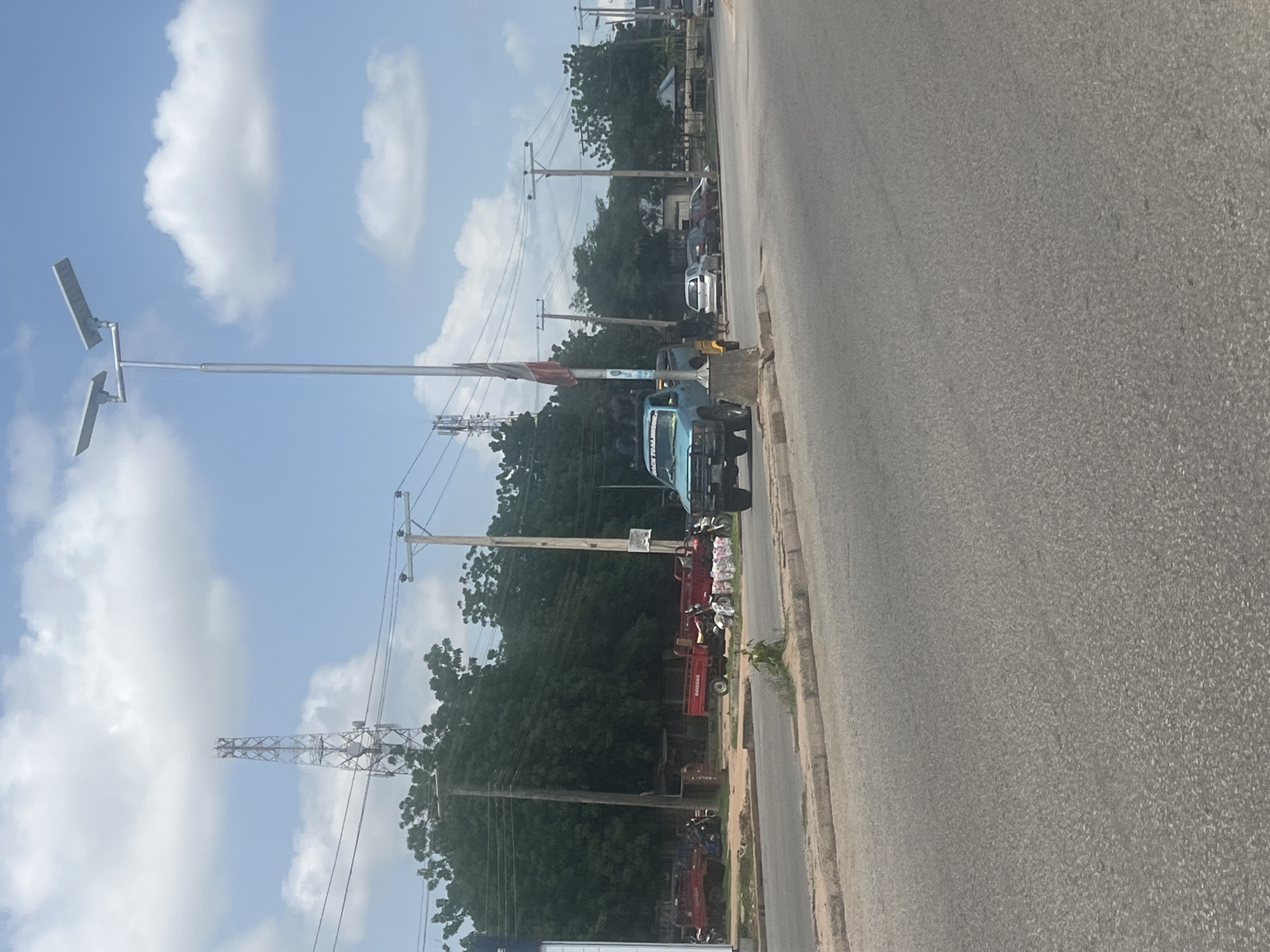 The image depicts a street scene in Jalingo, Nigeria. A bright blue pickup truck with a flag and several individuals in the bed is parked next to a lamppost. There are other vehicles, including red trucks and motorcycles, parked on the side of the road. In the background, trees line the street, and communication towers and utility poles are visible against a partly cloudy sky. The road is paved, with a curb separating it from the roadside.
