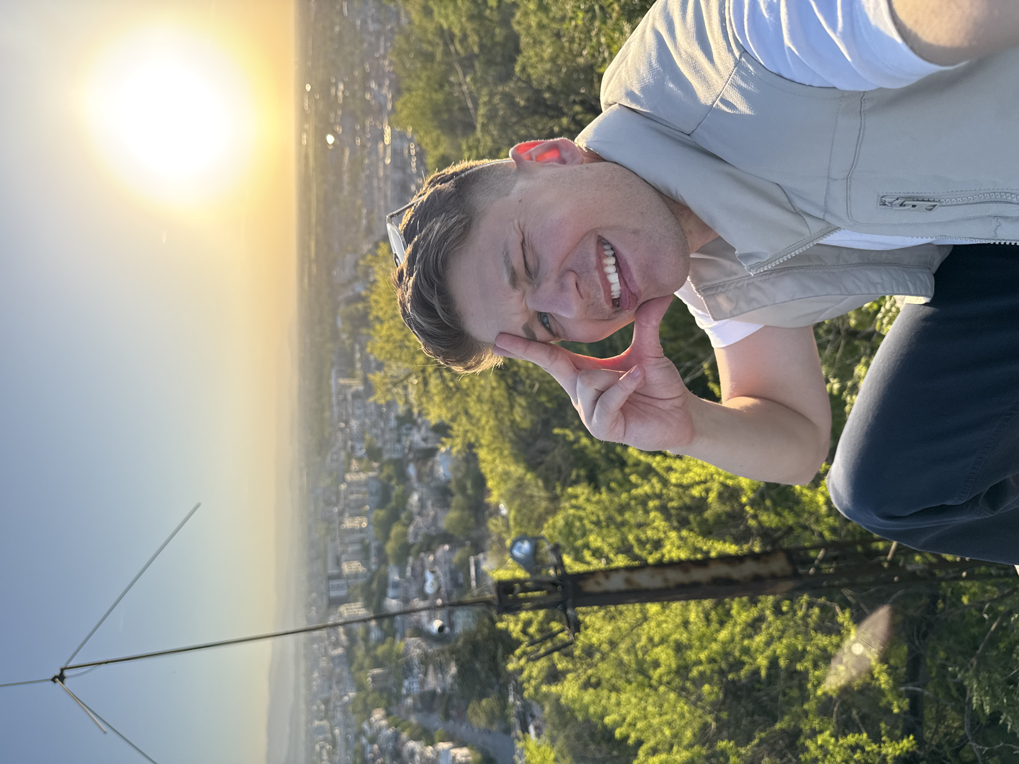 The image, rotated 90 degrees clockwise, captures a cheerful young man taking a selfie from a high vantage point overlooking a city during the golden hour. The man, positioned in the middle-right, winks and smiles broadly, showing teeth, while making a peace sign with his right hand. He wears a white t-shirt and a light beige vest, with sunglasses perched on his head.
The bright, warm sun dominates the upper-left corner of the sky, indicating either sunrise or sunset and casting a strong backlight that outlines the man's features. The sky transitions from a brilliant golden yellow around the sun to a hazy blue in the distance. Below him, a sprawling city, identifiable as Plovdiv, Bulgaria, stretches out, partially obscured by atmospheric haze. Lush green trees and vegetation cover the foreground and mid-ground, suggesting the viewpoint is from a hill or mountain. A metal antenna or pole structure is visible in the lower-right, blending with the greenery. The man's expression conveys happiness and playfulness. There is no visible text.