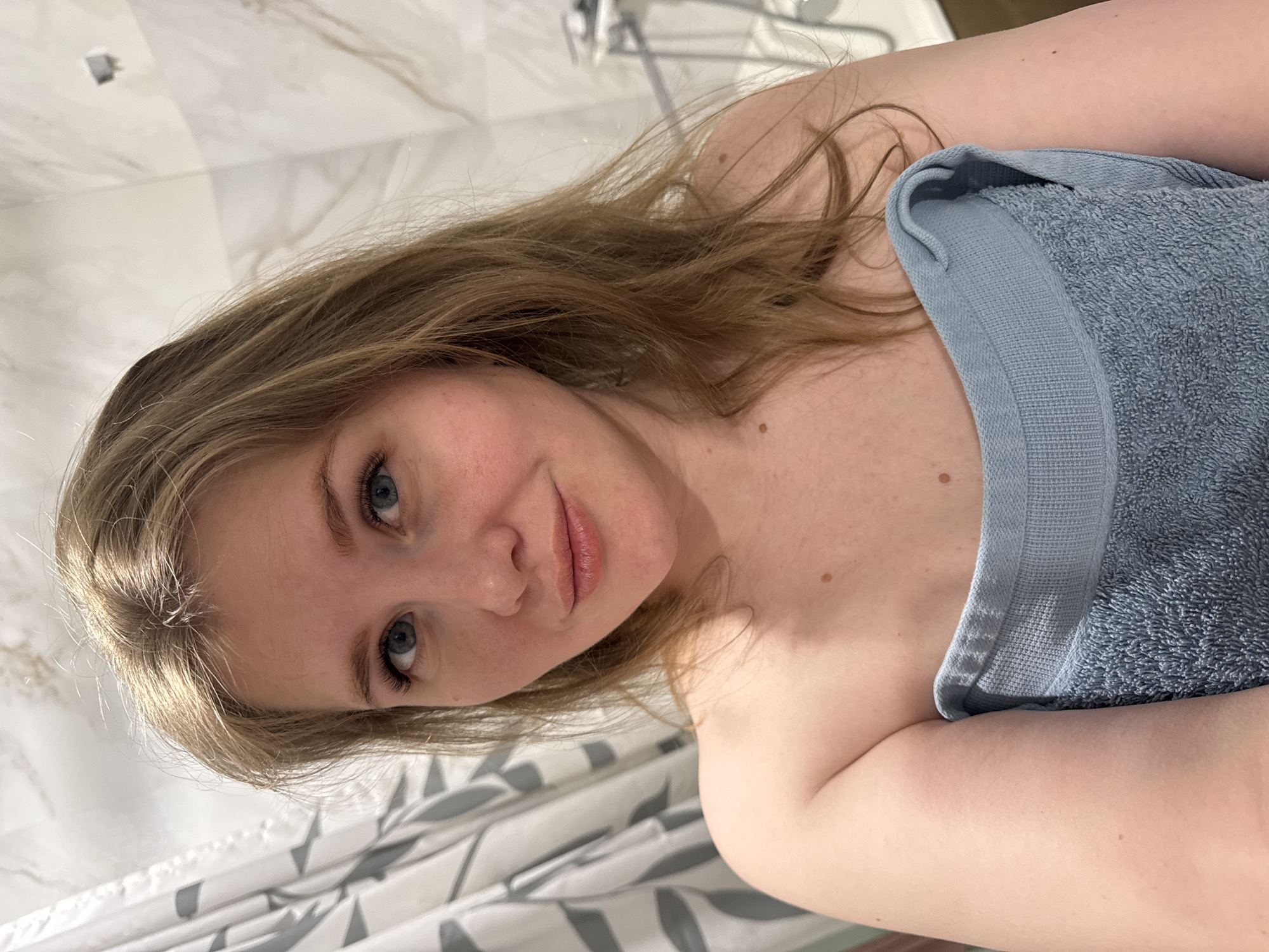 A young woman with light brown hair and blue eyes poses in a bathroom. She is partially covered by a light blue towel. The background includes a shower and a shower curtain with a gray and white leaf pattern. The location is likely a private residence. The city is Ферма, Russia.