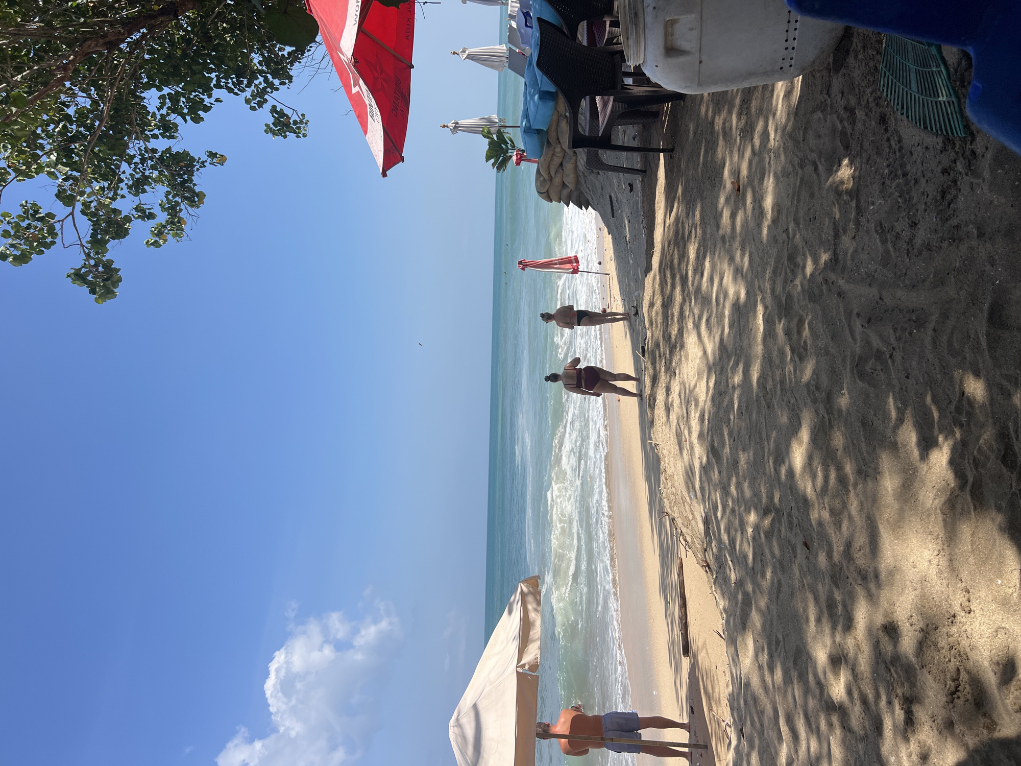 A wide shot captures a sunny day at Kuta Beach, Bali, Indonesia. The ocean is a vibrant turquoise, with moderate waves rolling onto the sandy shore. Two people are standing near the water's edge, facing the sea. One person, wearing a dark swimsuit, is closer to the camera. The other, in a reddish-brown swimsuit, is slightly further away.
To the left, a large, light-colored beach umbrella provides shade for a man standing nearby, facing the ocean and holding a cell phone. The sand is a warm golden color, with shadows cast by an unseen tree in the foreground, suggesting it's mid-day.
To the right, a red umbrella shades an area with blue cushions, tables, and chairs. Sandbags are stacked behind the seating area, forming a low wall. Several white beach umbrellas are visible further down the beach. The scene conveys a relaxed atmosphere at a popular tourist destination.