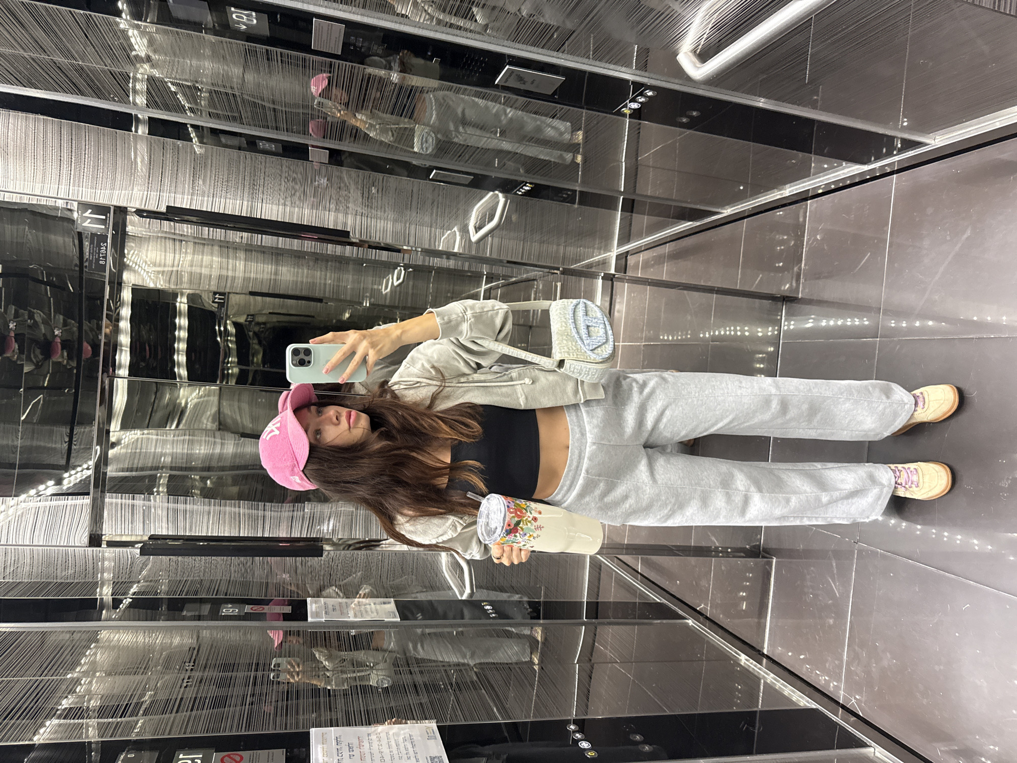 A young woman is taking a selfie in a modern, highly reflective elevator in Ramat Gan, Israel. She is seen holding a light blue iPhone to capture her reflection, with her gaze directed towards the camera. She wears a casual outfit consisting of a pink baseball cap, a light grey zip-up hoodie over a black crop top, and matching light grey sweatpants, paired with light-colored sneakers that have pink laces. In her other hand, she holds a large, light-colored travel mug adorned with a colorful floral pattern and a straw. A small light blue quilted shoulder bag, featuring a prominent H-like logo, hangs from her right shoulder.
The elevator's interior is characterized by dark, glossy, and textured walls that create multiple reflections of the woman and the surrounding space, giving a sense of depth and intricate patterns. The floor and ceiling also appear dark and reflective, contributing to the sleek, contemporary design. Artificial lighting illuminates the confined space, making the time of day undiscernible. The woman maintains a neutral, composed expression. Visible on the reflective surfaces are various signs and indicators, some displaying Hebrew text, such as safety instructions or facility information, and reflected floor numbers, like "11" and "19", further situating the scene within its specified location.