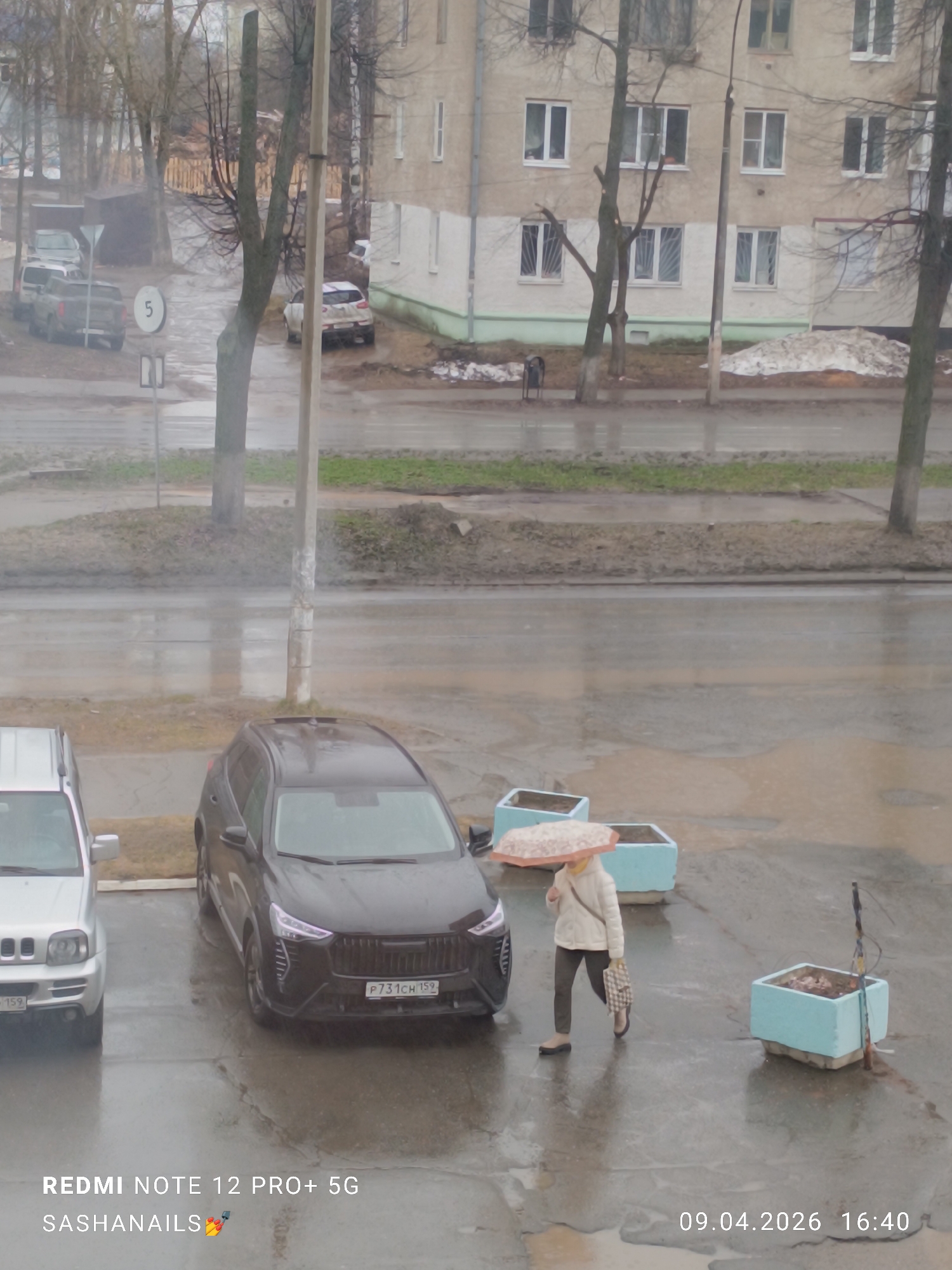 The image shows a rainy day in Chaikovsky, Russia. The timestamp indicates April 9, 2026, 16:40. It appears to be a residential or commercial area with multi-story buildings in the background. Several cars are parked on the wet pavement, including a black Haval SUV (registration P731CH159) and a silver SUV. A woman is walking with an umbrella and a bag. There are light blue concrete planters near the walking woman.