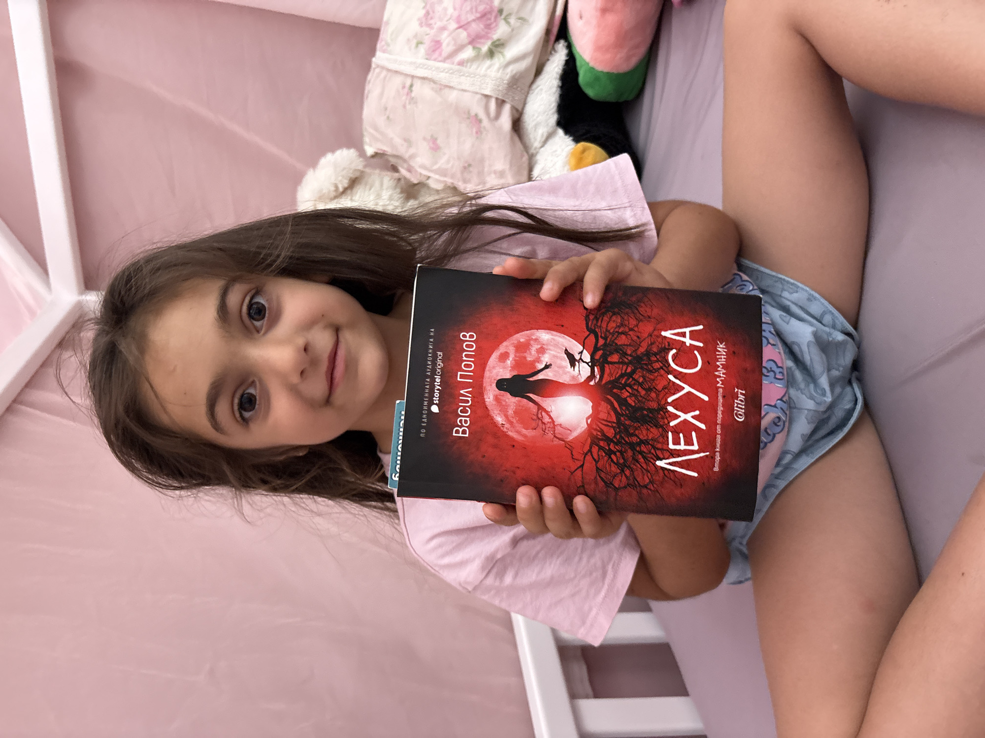 A young girl, with long brown hair and dark eyes, lies in bed, holding a book. The book's cover is predominantly red, featuring a silhouette of a woman against a large, pale moon, surrounded by bare, dark trees. The title, "ЛЕХУСА," is prominently displayed in white lettering. Smaller text on the book's spine indicates "Васил Попов," the author, and "storytel original." The girl is wearing a pink t-shirt and blue patterned shorts. She appears to be in a bedroom, with soft pink bedding and pillows behind her. The lighting suggests it could be daytime, possibly late afternoon. The girl's expression is pleasant, with a slight smile. A white crib frame is visible on the right side of the image. The overall scene conveys a sense of quiet relaxation and engagement with reading.