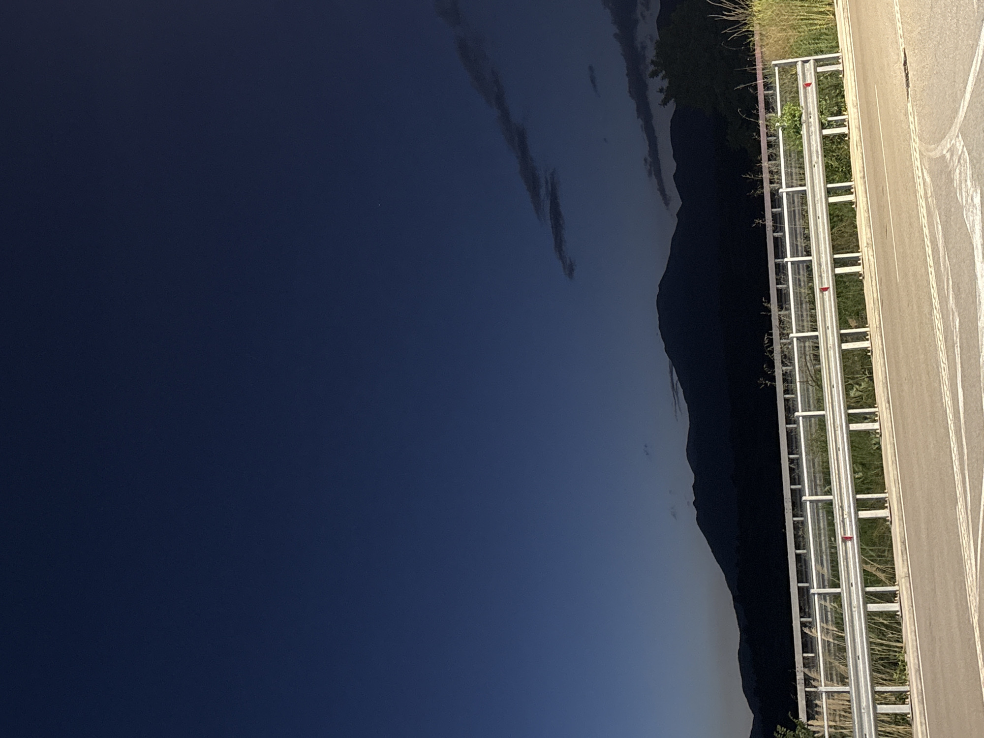 The image depicts a scene near Chuchuligovo, Bulgaria at dusk or night. To the left is a dark blue sky with scattered clouds. A silhouette of a mountain range extends along the right side of the sky. A roadside with a guardrail runs vertically on the right. The guardrail is white and metallic, with some vegetation growing around it. An asphalt road is visible to the right of the guardrail, marked with white lane lines.