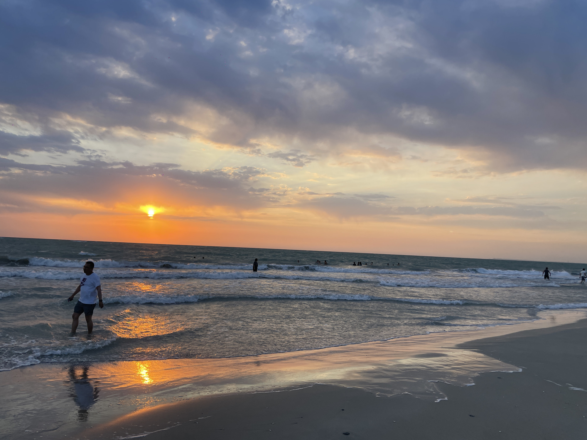 A man wearing a white t-shirt and dark shorts stands in the shallow water of a beach, facing the ocean and the setting sun. The sun, a bright orange disc, is low on the horizon, casting a warm glow across the sky, which is filled with dramatic clouds ranging from deep blue to pale orange and yellow. Waves gently lap at the shore, creating a reflective surface on the wet sand. In the background, several other people are visible in the water, some swimming or wading. The beach itself is sandy, with the wet sand in the foreground reflecting the colors of the sunset. The scene depicts a serene evening at the coast. The location is identified as Unknown, Kazakhstan.