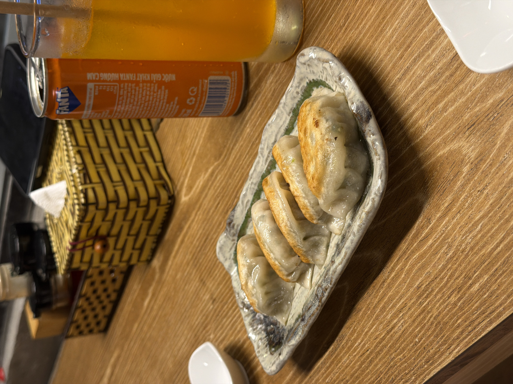 The image captures a close-up, slightly overhead view of a meal setting on a light brown wooden table with prominent grain. The primary focus is a ceramic, irregularly shaped plate holding five pan-fried dumplings (gyoza). The dumplings have a golden-brown, crispy bottom and a soft, pale top, indicating they are cooked.
To the upper left of the plate, a tall glass filled with an orange beverage, likely orange juice or a soft drink, stands with a straw. Partially visible behind the glass is an orange Fanta can. The Fanta can clearly displays its logo and Vietnamese text: "NƯỚC GIẢI KHÁT FANTA HƯƠNG CAM," which translates to "FANTA ORANGE FLAVOR SOFT DRINK." Nutritional information and a barcode are also visible on the can.
On the left side of the table, a woven yellow-brown tissue box holder with a checkered pattern is present. A small, white, tear-drop shaped dish, likely for dipping sauce, is positioned in the lower center foreground.
The scene suggests a casual dining experience, possibly in a restaurant or cafe in Da Nang, Vietnam, given the language on the Fanta can. The lighting indicates an indoor setting. No people are visible.