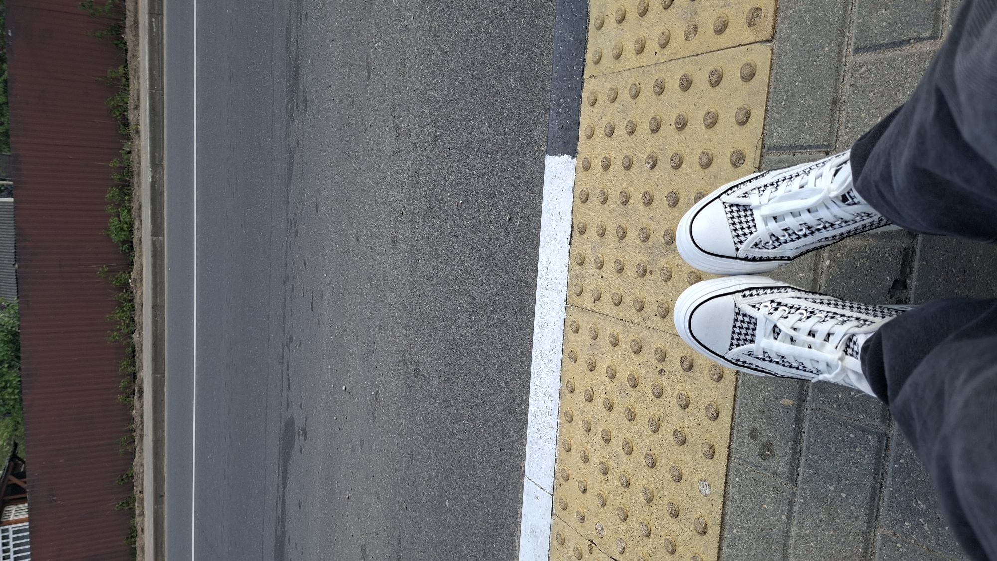 The image shows a view from above looking down at a person's feet standing on a sidewalk. The person is wearing houndstooth patterned sneakers and grey jeans. The sidewalk is made of two types of paving: one section is yellow with raised circular bumps, likely for tactile paving to assist visually impaired individuals, and the other is grey. To the left of the sidewalk is an asphalt road with a painted white line marking the edge. Beyond the road, a rusty corrugated metal fence or wall can be seen, partially obscured by greenery. The environment is typical of a city street. The presence of tactile paving suggests a pedestrian crossing. Without any additional information or features unique to that location, it is impossible to verify with certainty if the image depicts the city of Смалявічы, Belarus.