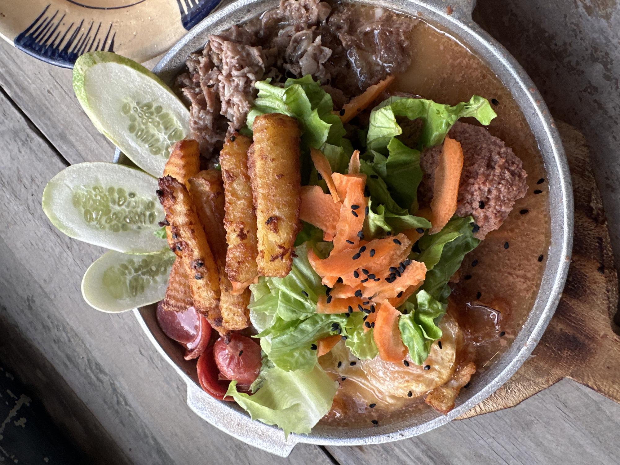 A close-up, overhead shot captures a sizzling platter of food served in a round, metallic dish with a wooden handle, placed on a weathered wooden surface. The dish contains a variety of ingredients: several thick-cut, golden-brown french fries are stacked in the center, nestled amongst crisp green lettuce leaves and shredded carrots. To the left, slices of red sausage and a small, pink patty are visible, partially submerged in a light brown sauce. The right side of the platter is filled with shredded meat, also in the sauce. Three oval-shaped slices of cucumber, their seeds clearly visible, adorn the top of the dish. Black sesame seeds are sprinkled over the carrots and lettuce, adding a touch of visual contrast. The overall presentation suggests a hearty and flavorful meal. The background reveals a rustic, wooden table, hinting at an outdoor or casual dining setting. There are no people present in the image. The provided location of Tây Hoa Lư Ward, Vietnam, is noted, but the image itself does not contain any specific visual cues to confirm or deny this precise location.