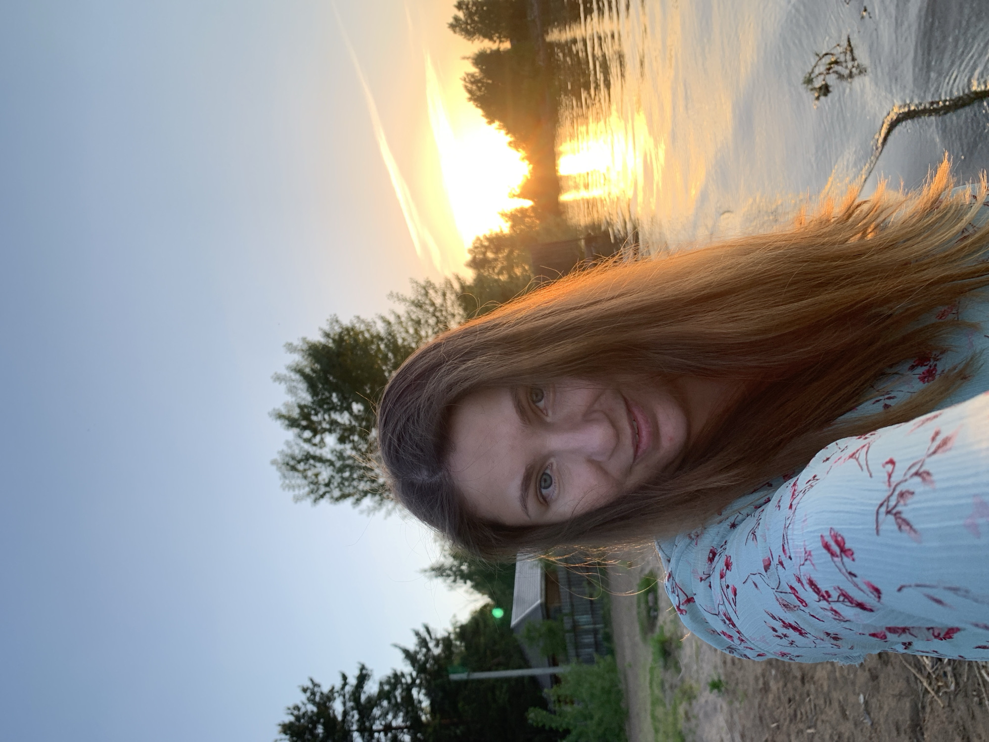 A woman with long brown hair and light eyes is taking a selfie at sunset. The sun is setting over a body of water, with reflections visible on the surface. Trees are visible in the background. The woman is wearing a light blue floral print dress. The location appears to be near a body of water with a wooded area, likely in the vicinity of Chaikovsky, Russia given that it's specified in the prompt. A building can be seen among the trees.