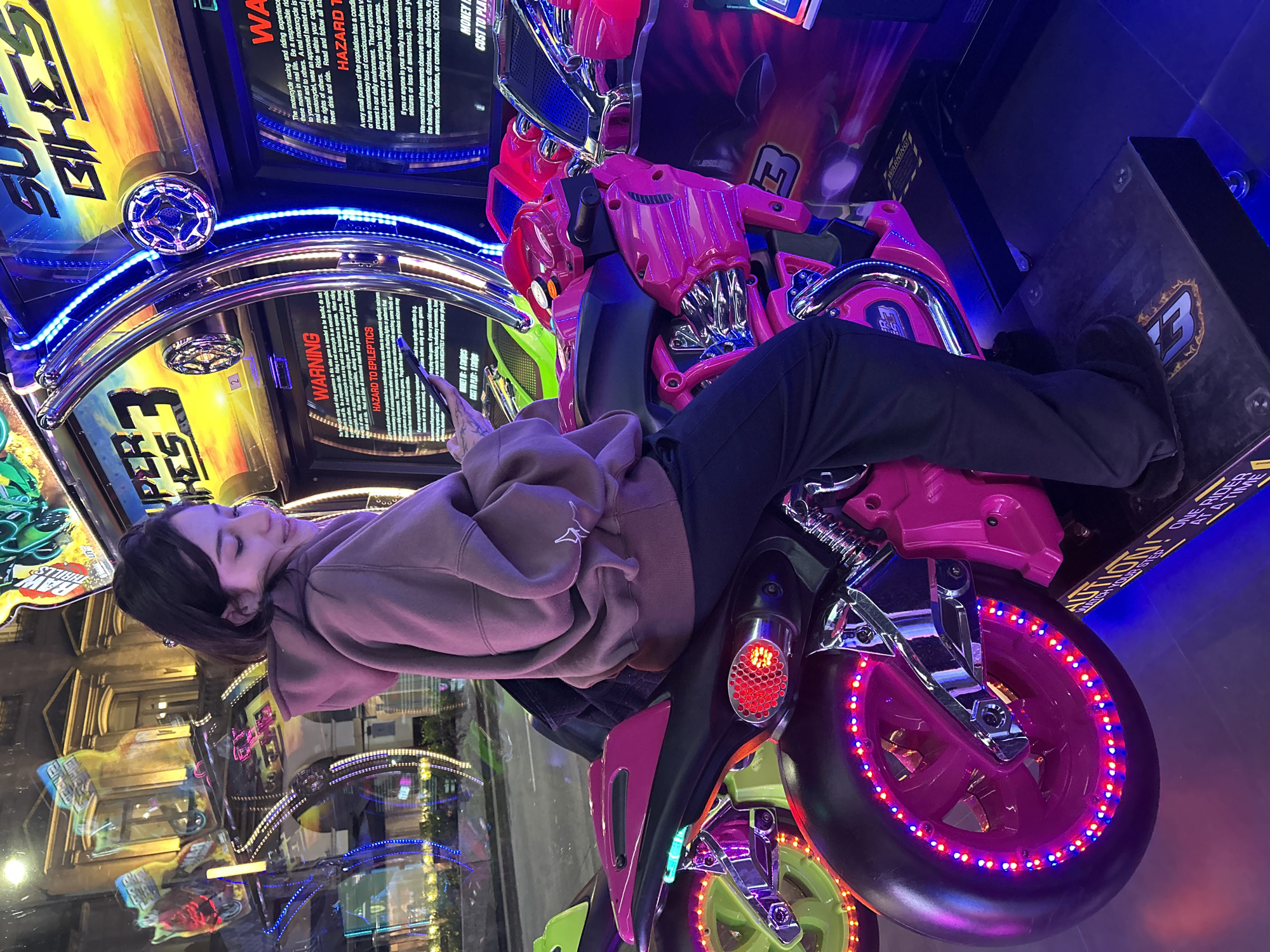 An eye-level shot captures a young woman seated on a bright pink and green motorcycle arcade racing game, "SUPER BIKES 3," within an arcade in Buenos Aires, Argentina. The indoor setting is dimly lit by ambient light, contrasting with the vibrant neon blue, pink, and green LEDs emanating from the arcade machine itself, particularly around the wheels and control panel. The woman, wearing a brown hoodie and black pants, appears relaxed, with her head gently tilted and eyes closed or cast downwards as she holds a smartphone.
The arcade motorcycle is prominently featured, boasting a glossy pink body, black seat, and neon green accents. Its wheels are adorned with strips of red and blue LED lights, creating a dynamic, glowing effect. Reflections on the large screen of the arcade game show other parts of the entertainment center, including what might be architectural elements in the background, hinting at the lively environment.
Multiple warning labels are visible on the machine's screens and frame. Text includes general "WARNING" messages, one specifically cautioning about "HAZARD TO EPILEPTICS" with detailed explanations about symptoms and consulting a physician. Other text advises on responsible riding, emphasizing that the game is not a substitute for real-life experience and recommending safety practices like wearing a helmet. Instructions for playing are also visible, stating "MONEY ON: O Swipes" and "COST TO PLAY: 1 Swipe." Additionally, a label warns, "WATCH YOUR STEP!" and "ONE RIDER AT A TIME." The overall atmosphere is one of leisure and entertainment.